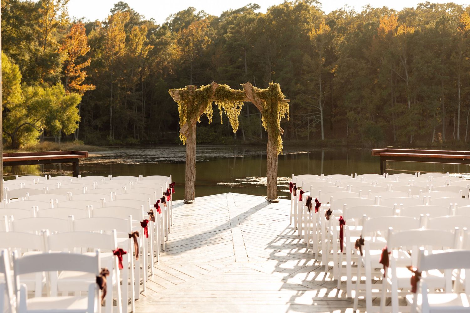 Outdoor wedding ceremony setup with white chairs and a decorated wooden arch overlooking a lake at sunset.