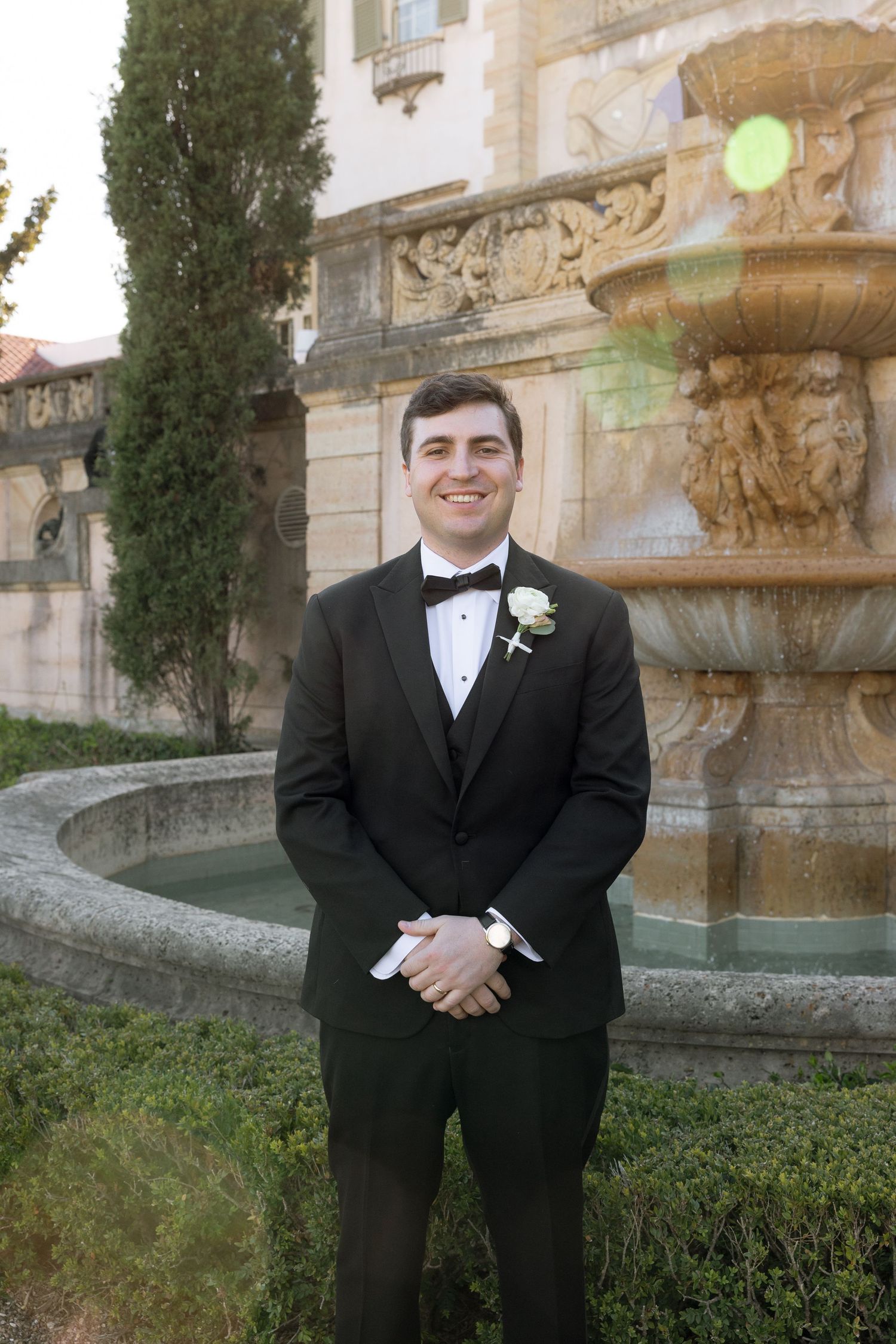 Smiling groom in black tuxedo standing by ornate stone fountain outdoors.