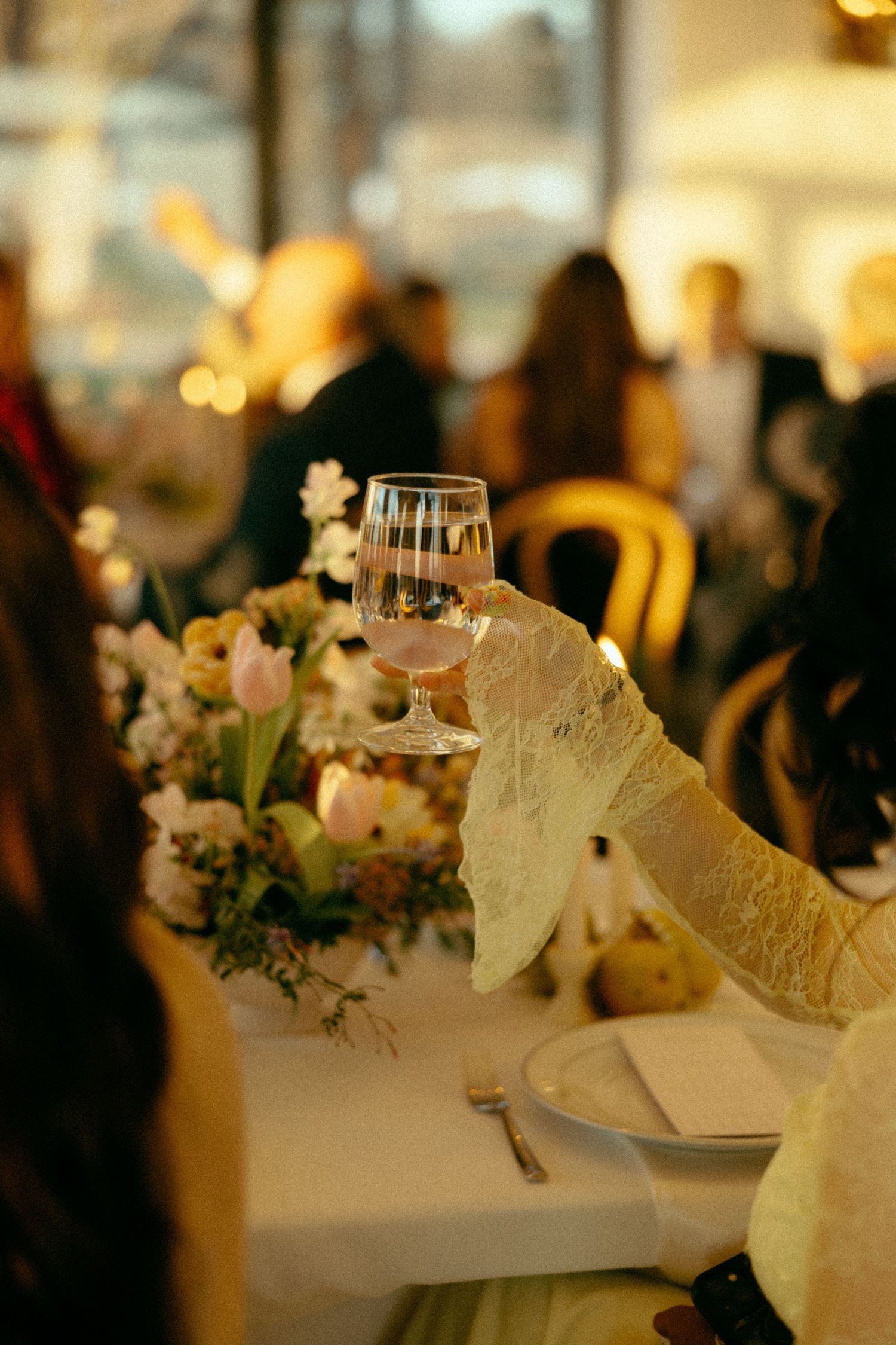 Elegant wedding reception toast with lace glove holding wine glass above floral centerpiece with tulips and candles.