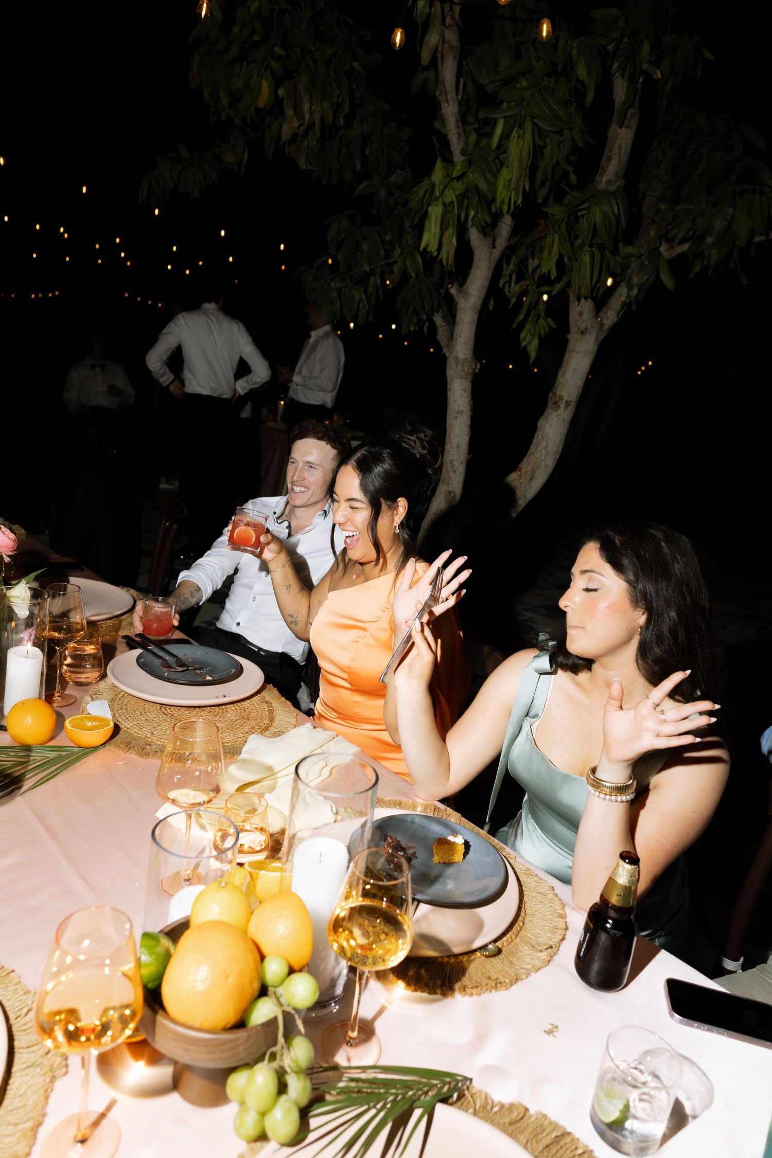 Guests in elegant attire laugh and chat at an outdoor evening wedding reception dinner table.