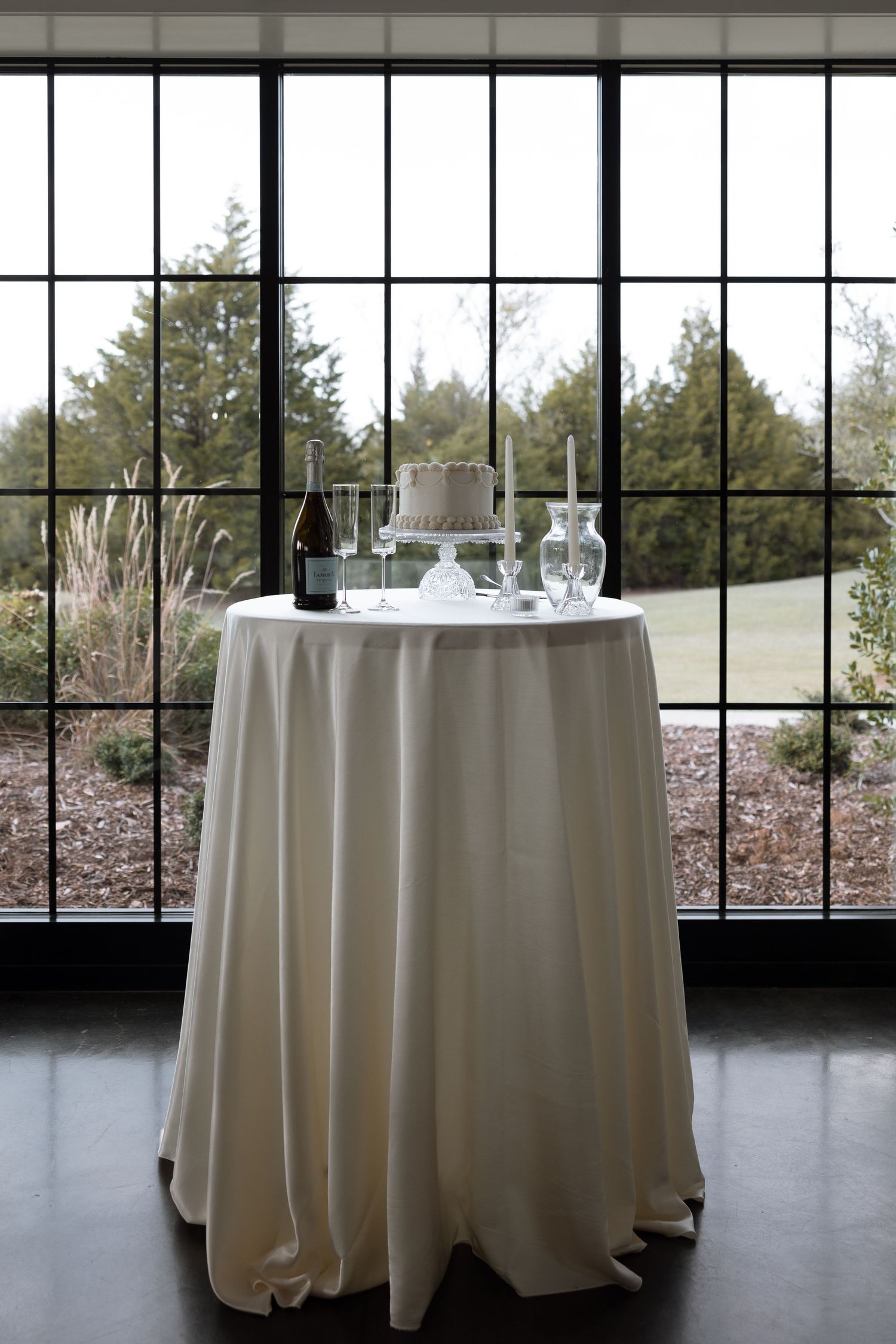 Round cocktail table with white tablecloth set against large windows overlooking trees.