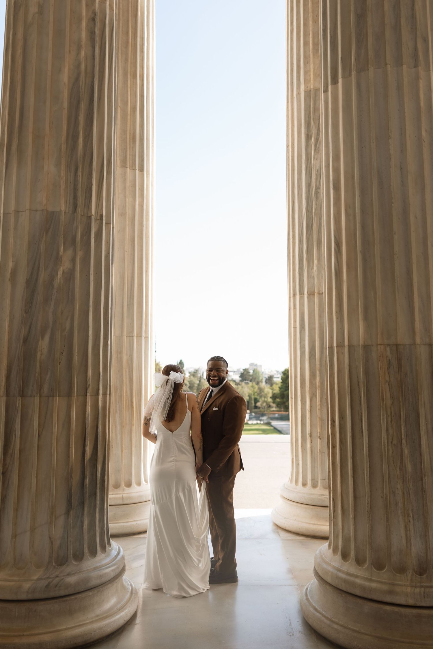 Couple poses between massive marble columns with bright sky in background.