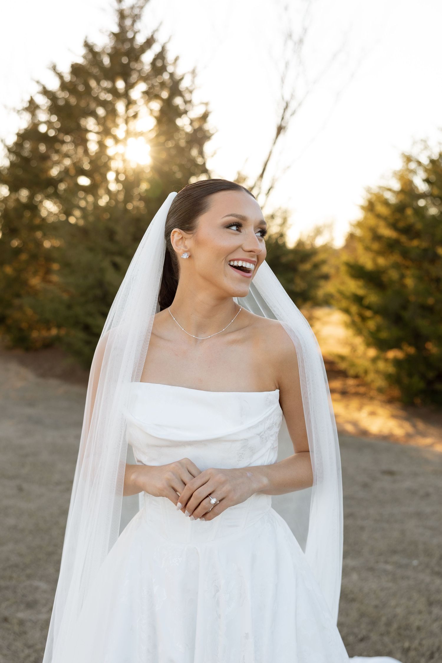 Joyful bride in strapless white dress and flowing veil poses outdoors at sunset with evergreen trees in background.
