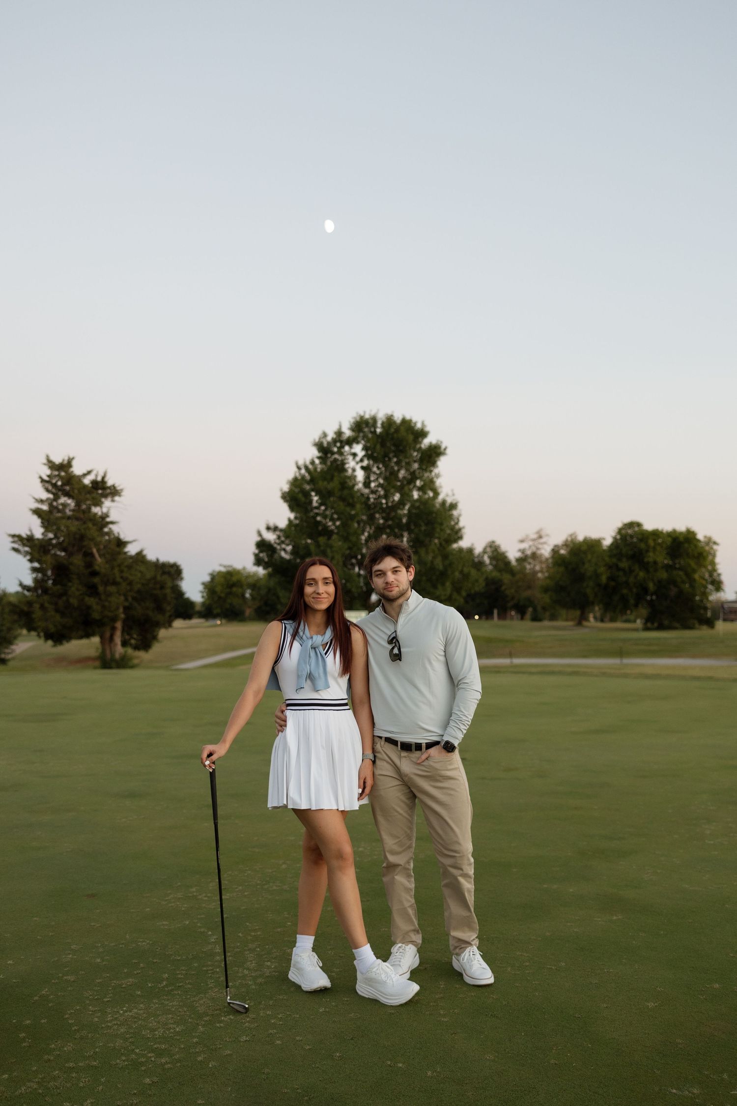 A couple poses with golf clubs on a lush green course during golden hour.