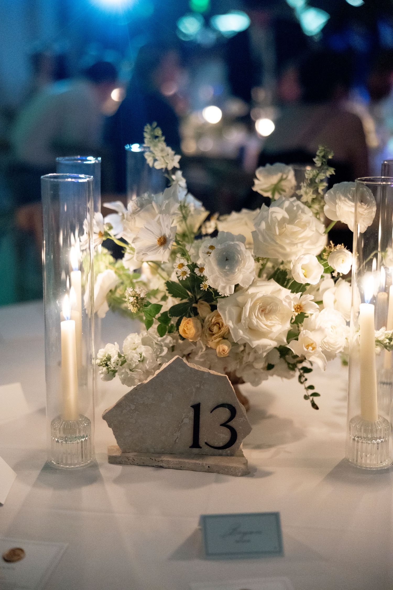 Elegant wedding table setting with white floral centerpieces, glowing candles, and table number 13 displayed on a stone holder.