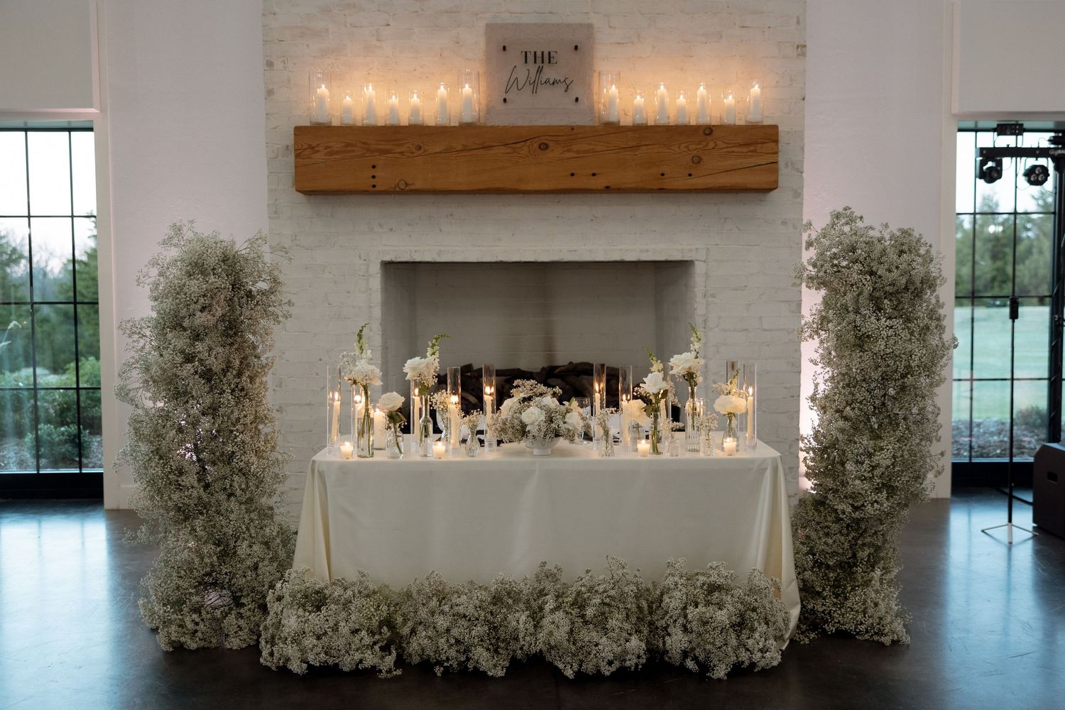Elegant white floral wedding reception table with candles and decor against a white brick fireplace.