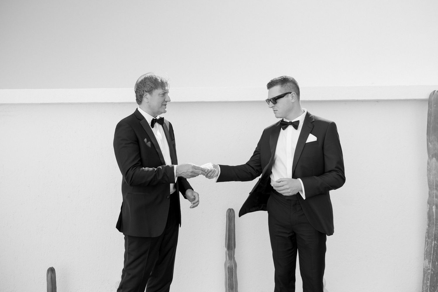 Two men in black tuxedos shake hands in black and white wedding photo indoors.