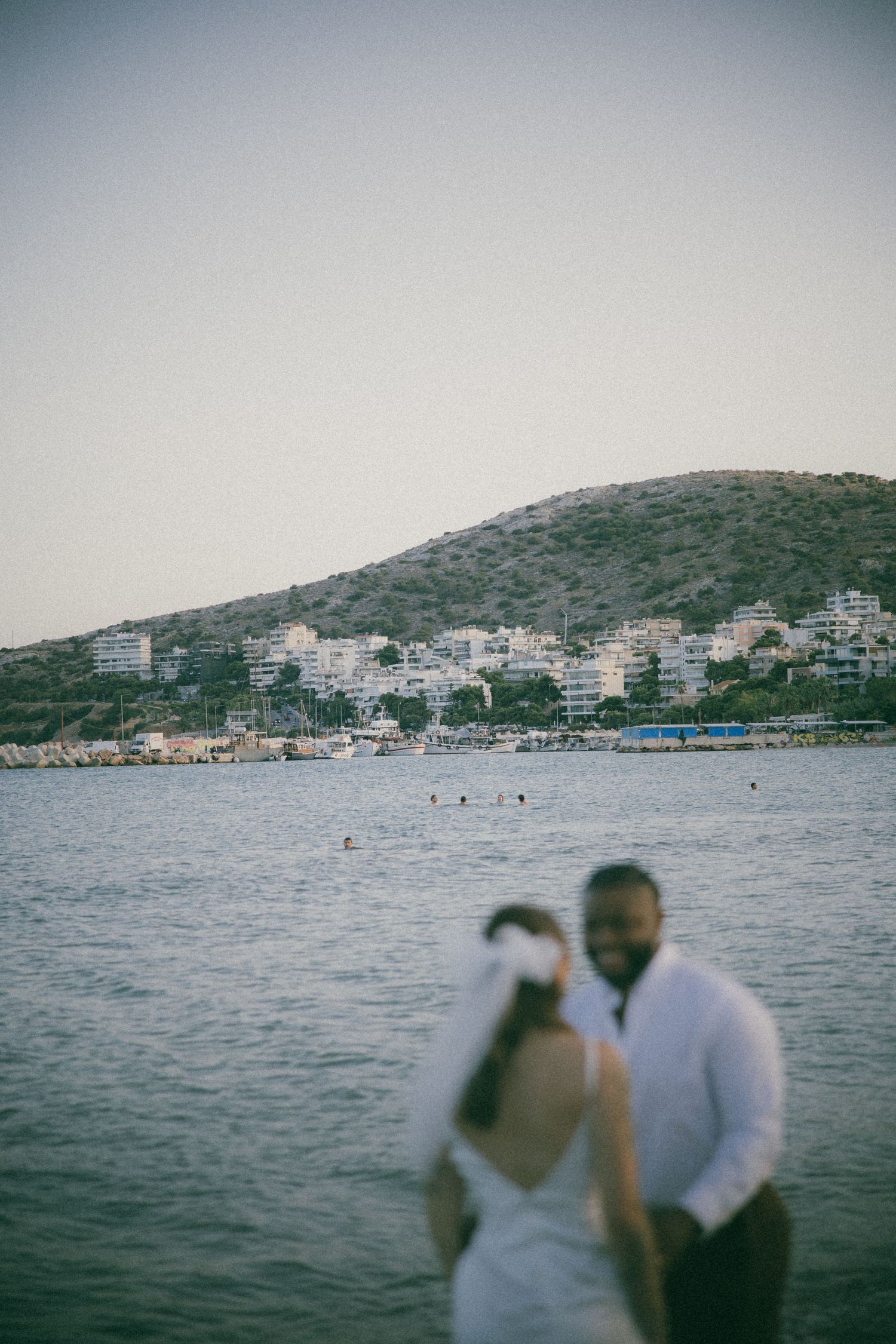 A romantic couple gazes at the Mediterranean seaside view with boats and buildings dotting the coastline.