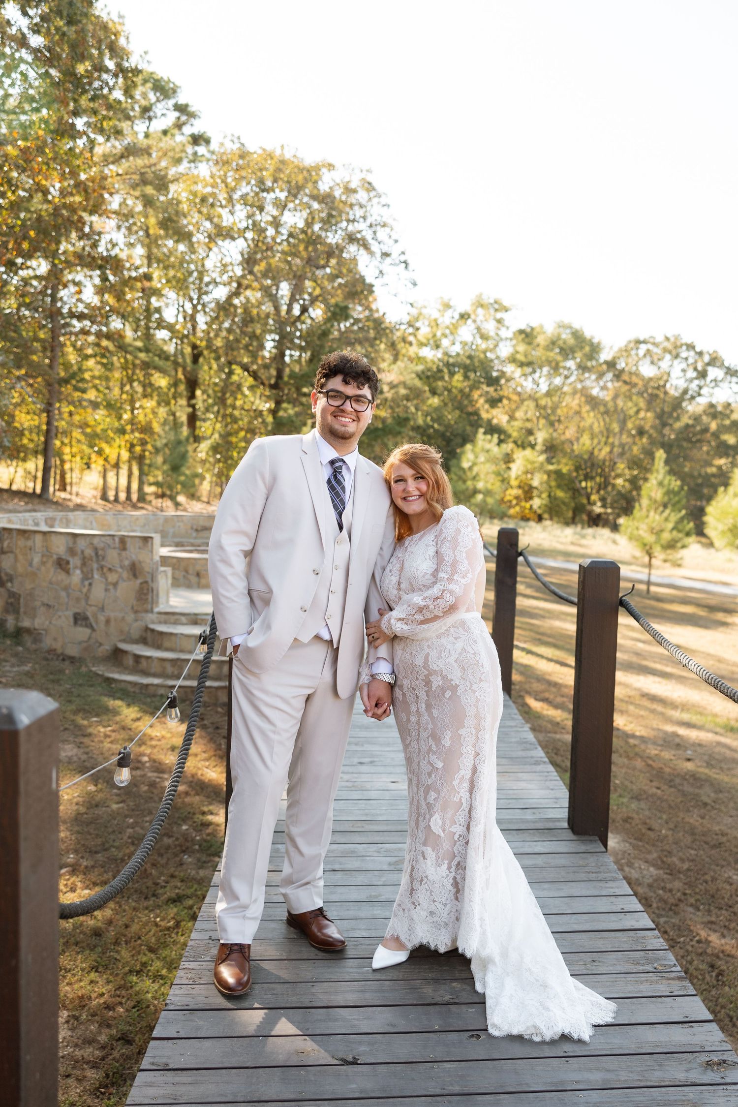 Newlyweds in white attire pose together on a rustic wooden walkway surrounded by fall trees.