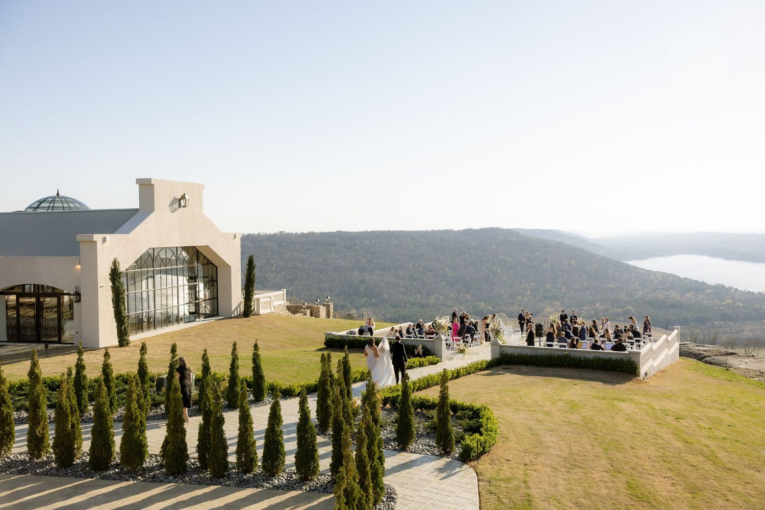 Elegant hilltop wedding venue with mountain views, manicured cypress trees, and guests gathered on a scenic outdoor terrace.