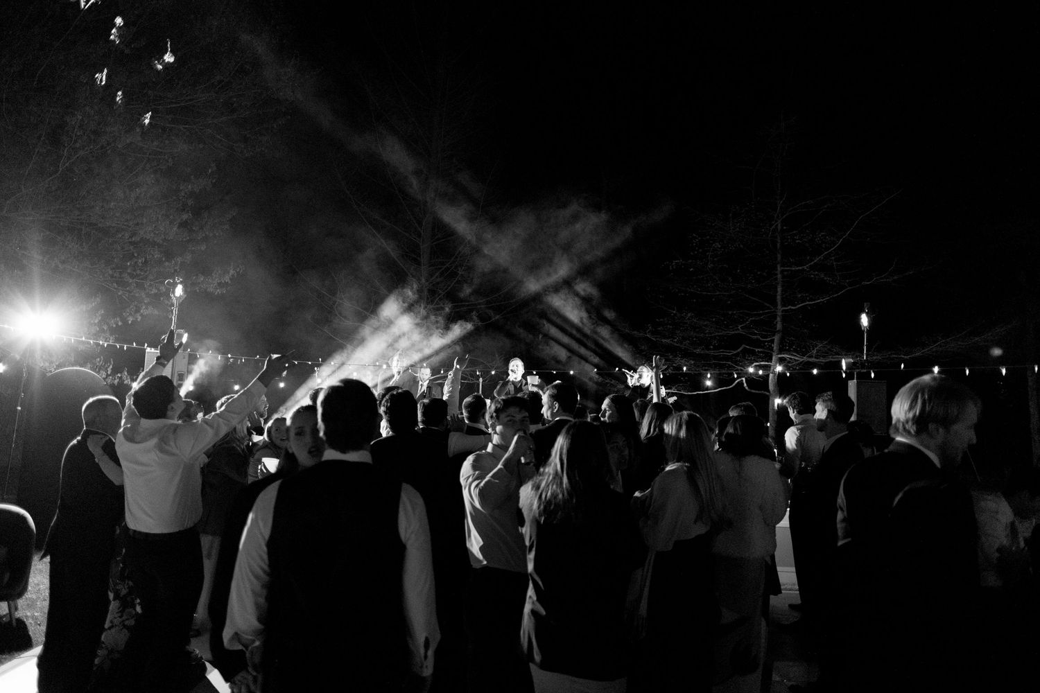 Black and white photo of wedding guests dancing and celebrating under dramatic stage lights at night.