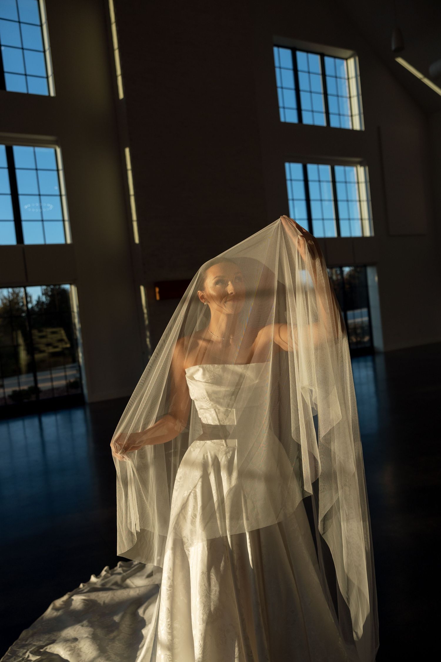 Ethereal silhouette of a bride covered in sheer veil against dramatic window lighting.