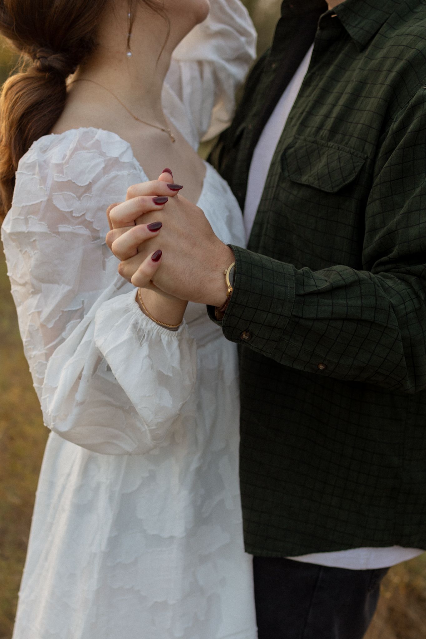 Close-up of clasped hands showing a white lace dress sleeve and green jacket in warm sunlight.