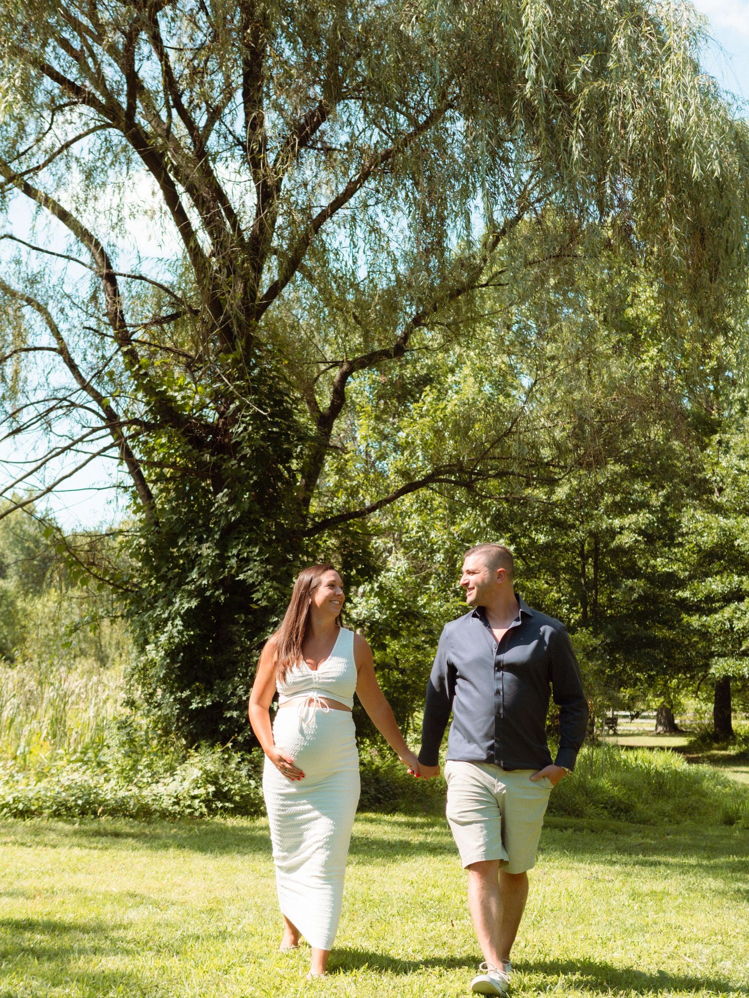 Pregnant couple walks hand-in-hand through lush green park under willow tree on sunny day.