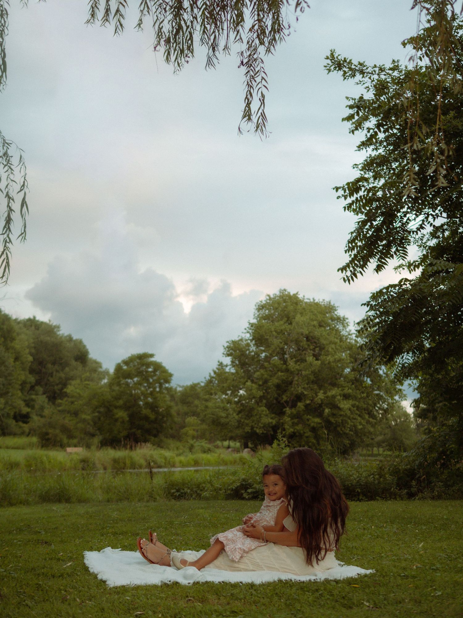 Mother sitting on blanket with toddler daughter in lush green park under willow tree at dusk.