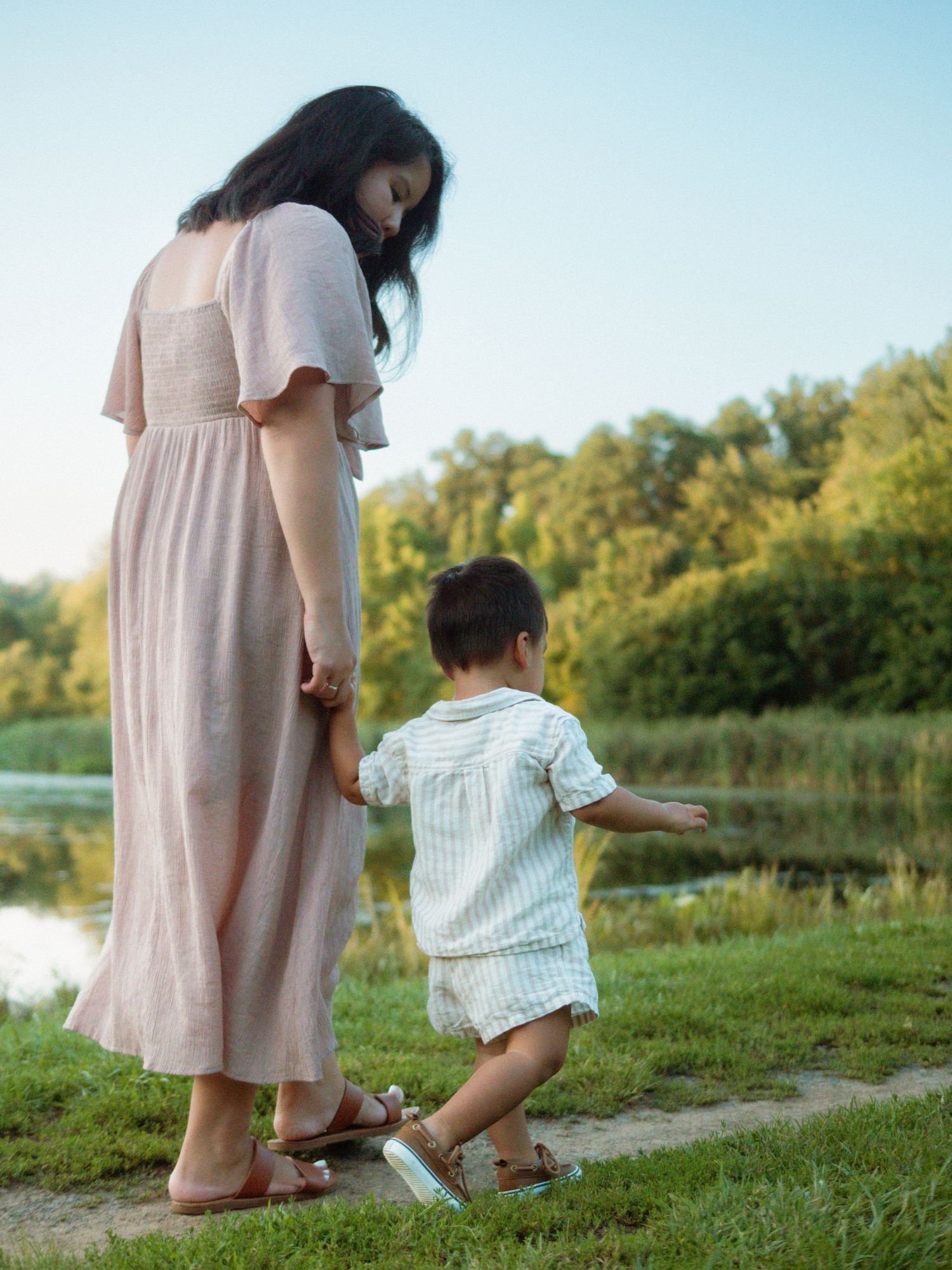 Mother holding toddler's hand by a peaceful lakeside on a sunny day.