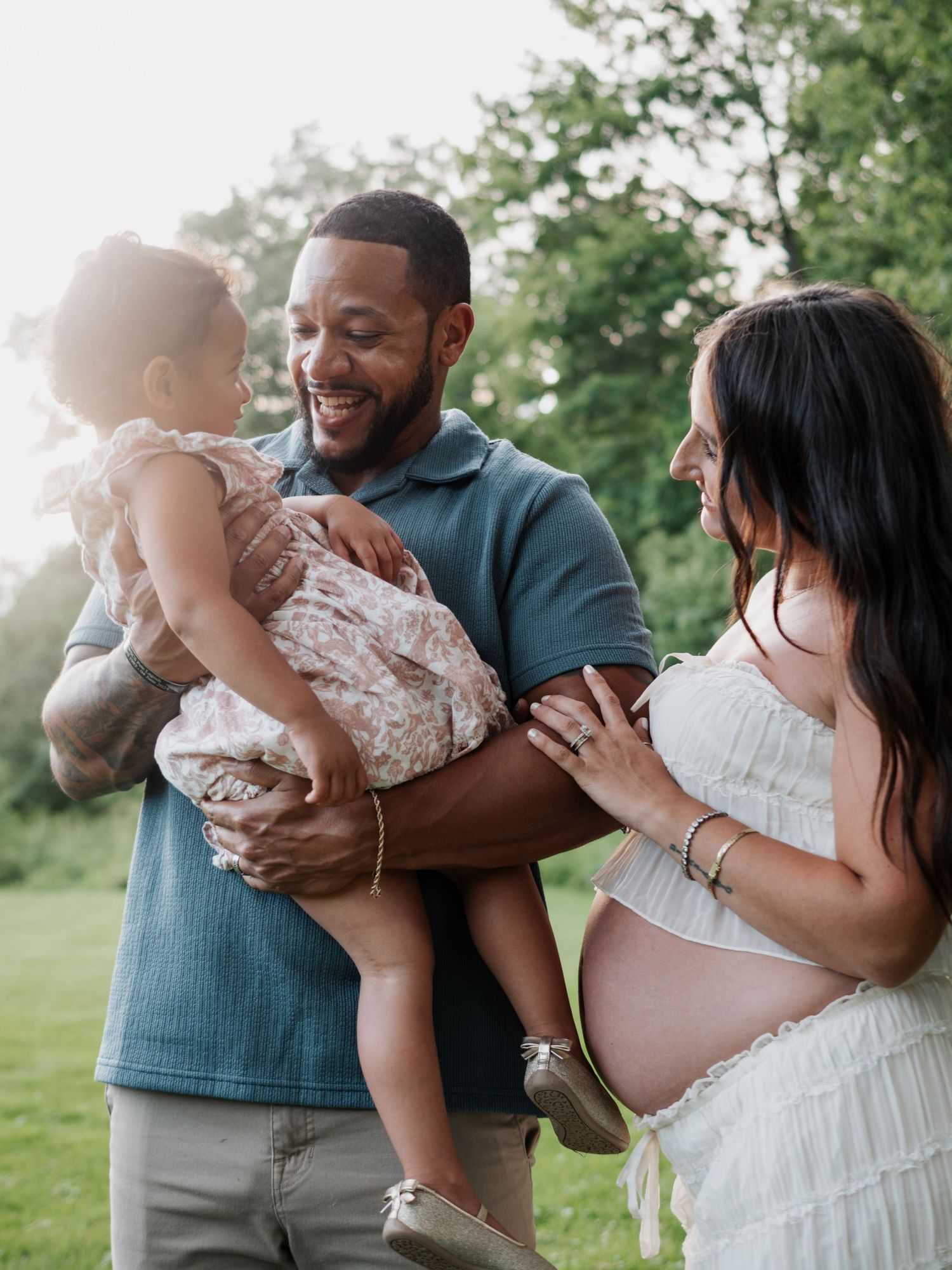 Smiling expectant parents share a joyful moment with their toddler daughter in a sunny green park.