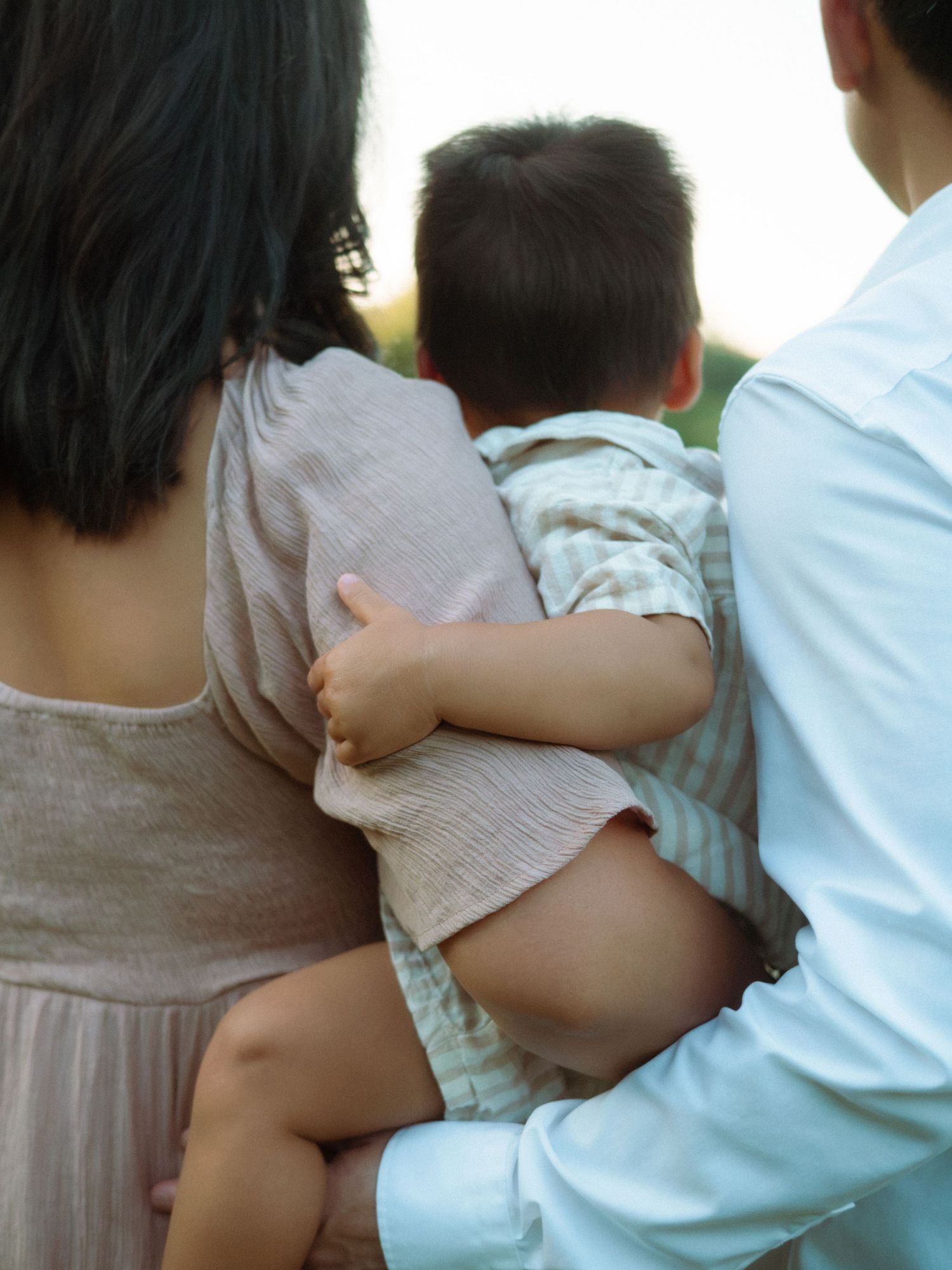 Close-up rear view of parents cradling their baby outdoors in soft evening light.
