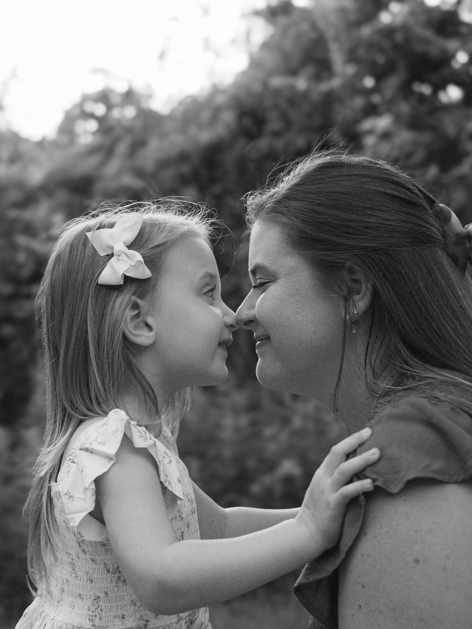 Black and white photo of mother and young daughter touching noses and smiling outdoors.