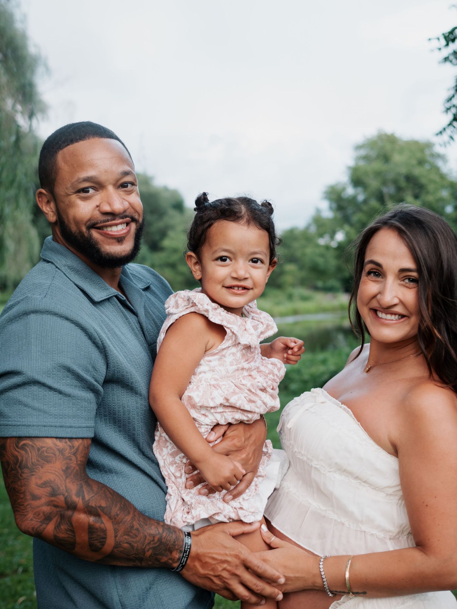 Smiling family portrait with tattooed father, pregnant mother, and toddler daughter outdoors.
