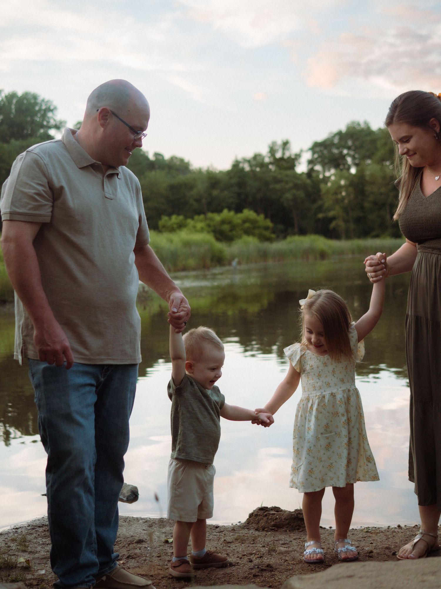 Family of four holding hands by a reflective lake at sunset in a serene outdoor setting.