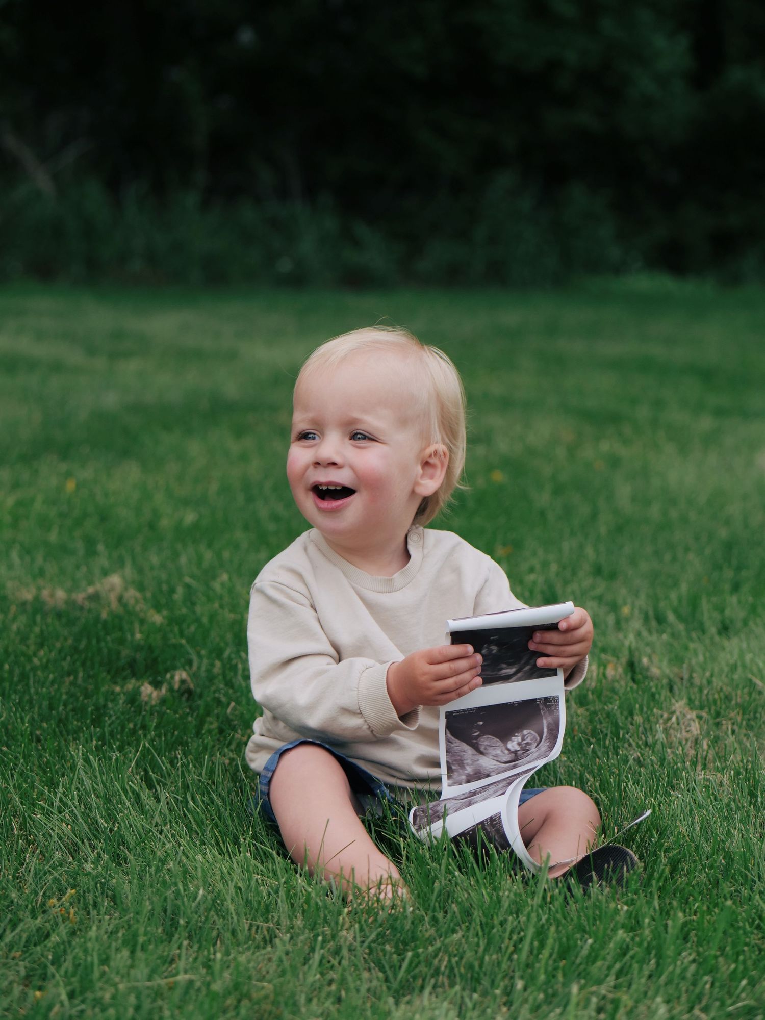 Happy laughing baby sitting on green grass holding ultrasound photos outdoors.