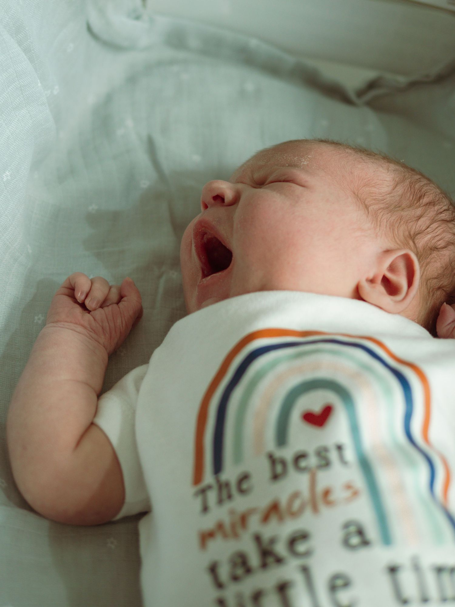 Newborn baby yawning in a white onesie reading the best miracles take a little time.