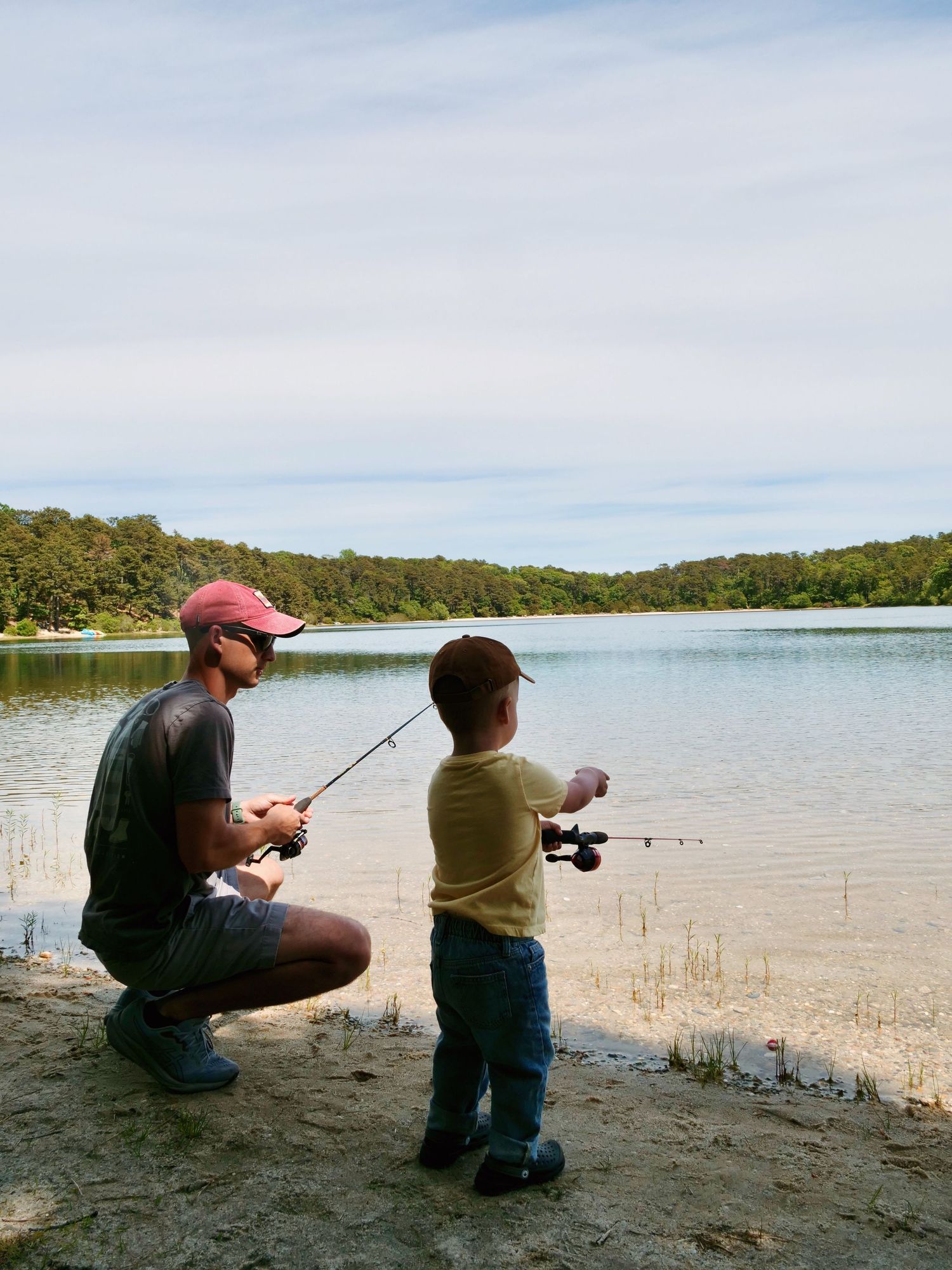 Father and young son fishing together at peaceful lakeside on a cloudy day.