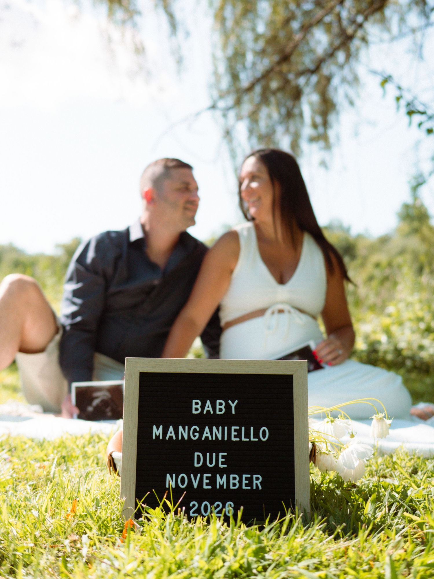 Baby Manganiello due November 2026 sign in foreground with expecting couple sitting behind it.