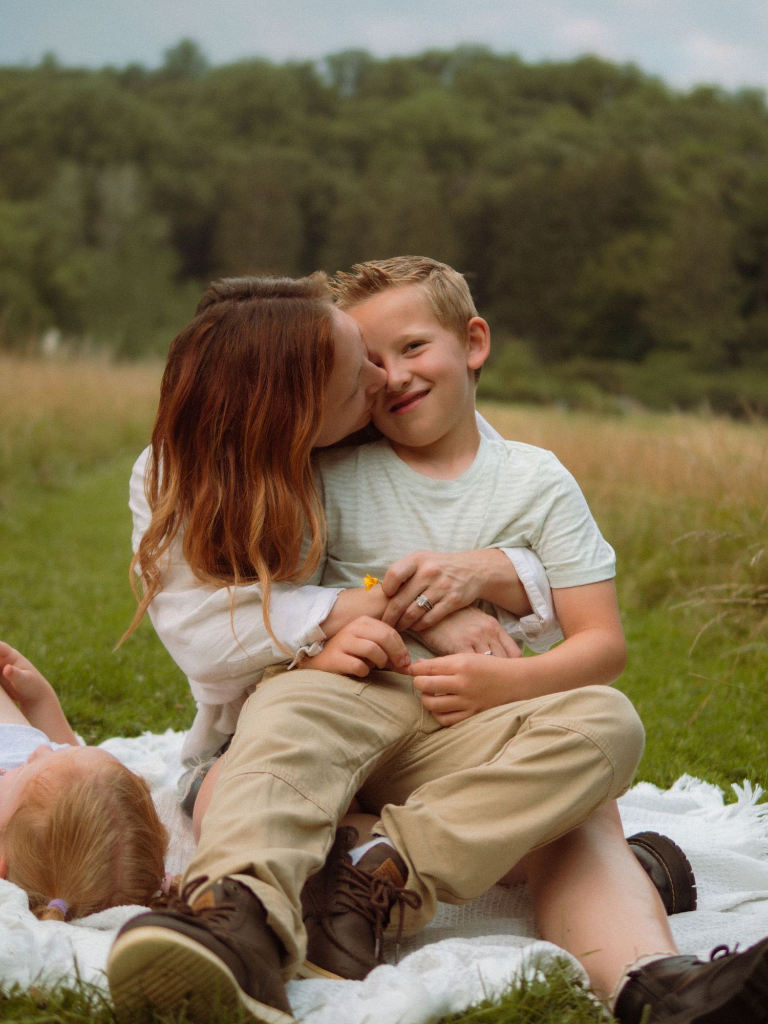 Mother kisses smiling young boy on cheek while sitting on white blanket in green meadow.