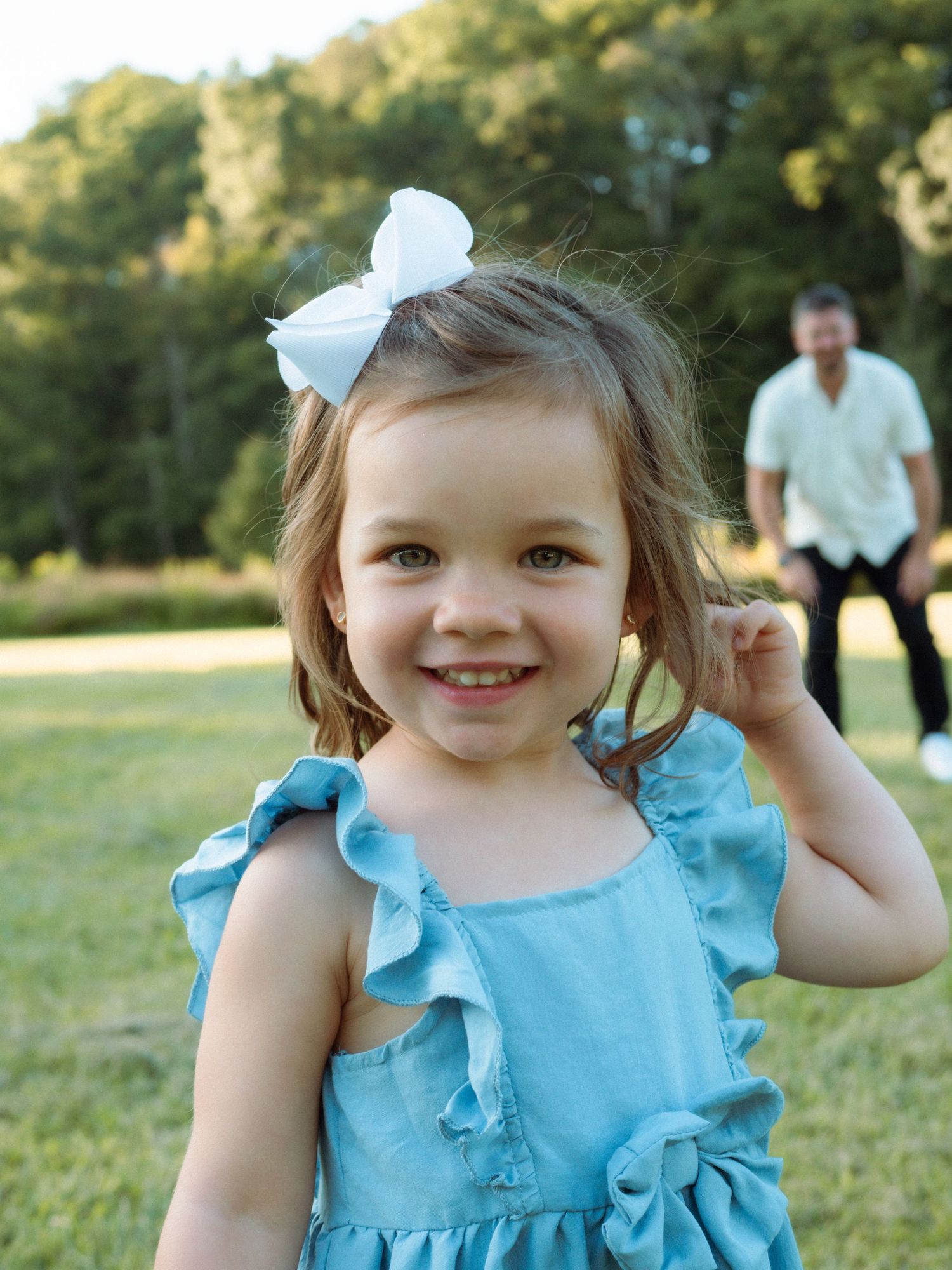 Adorable little girl in blue dress with white bow smiling in a park.