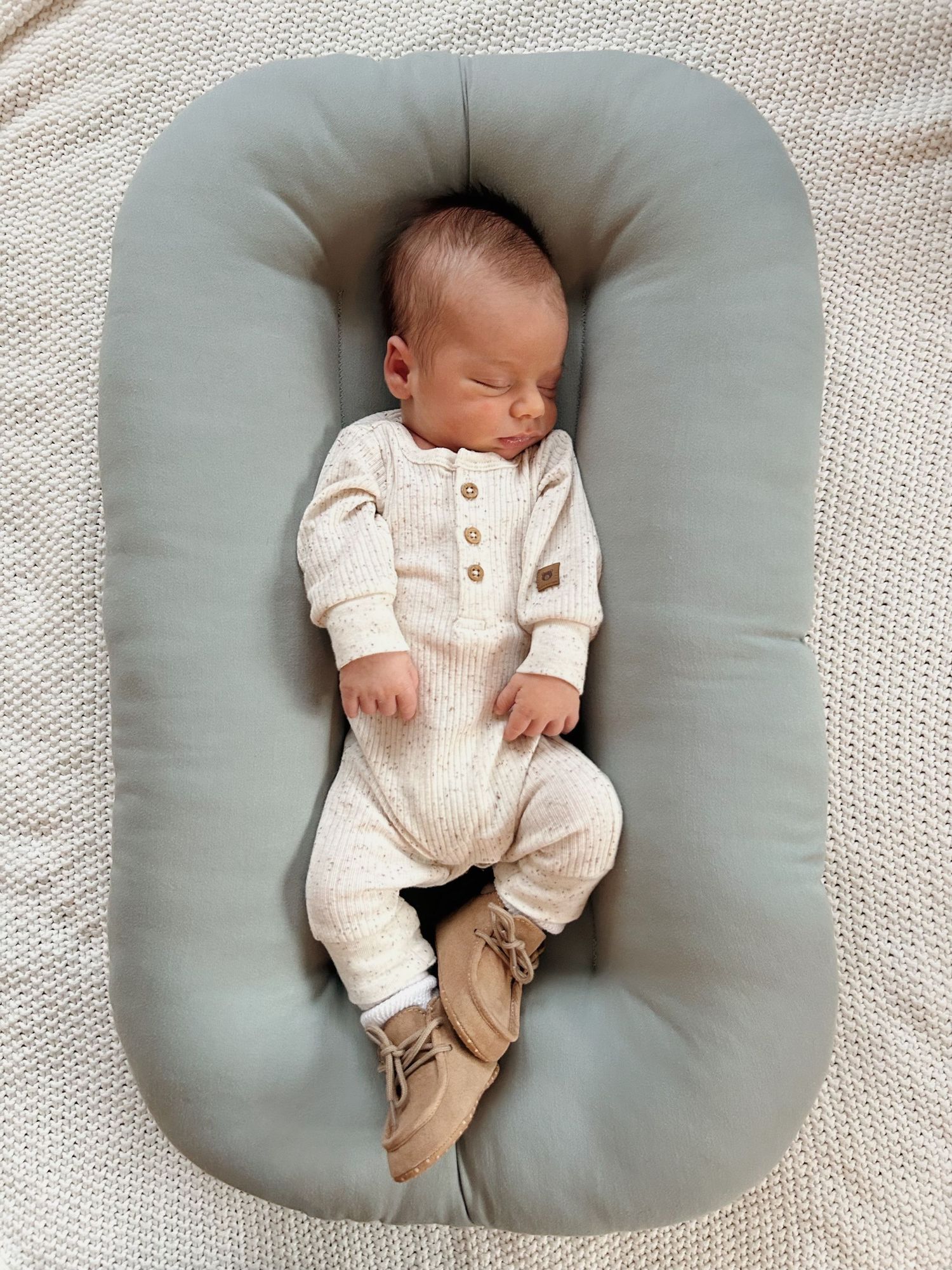 Newborn baby sleeping in a cream ribbed romper inside a sage green baby lounger.