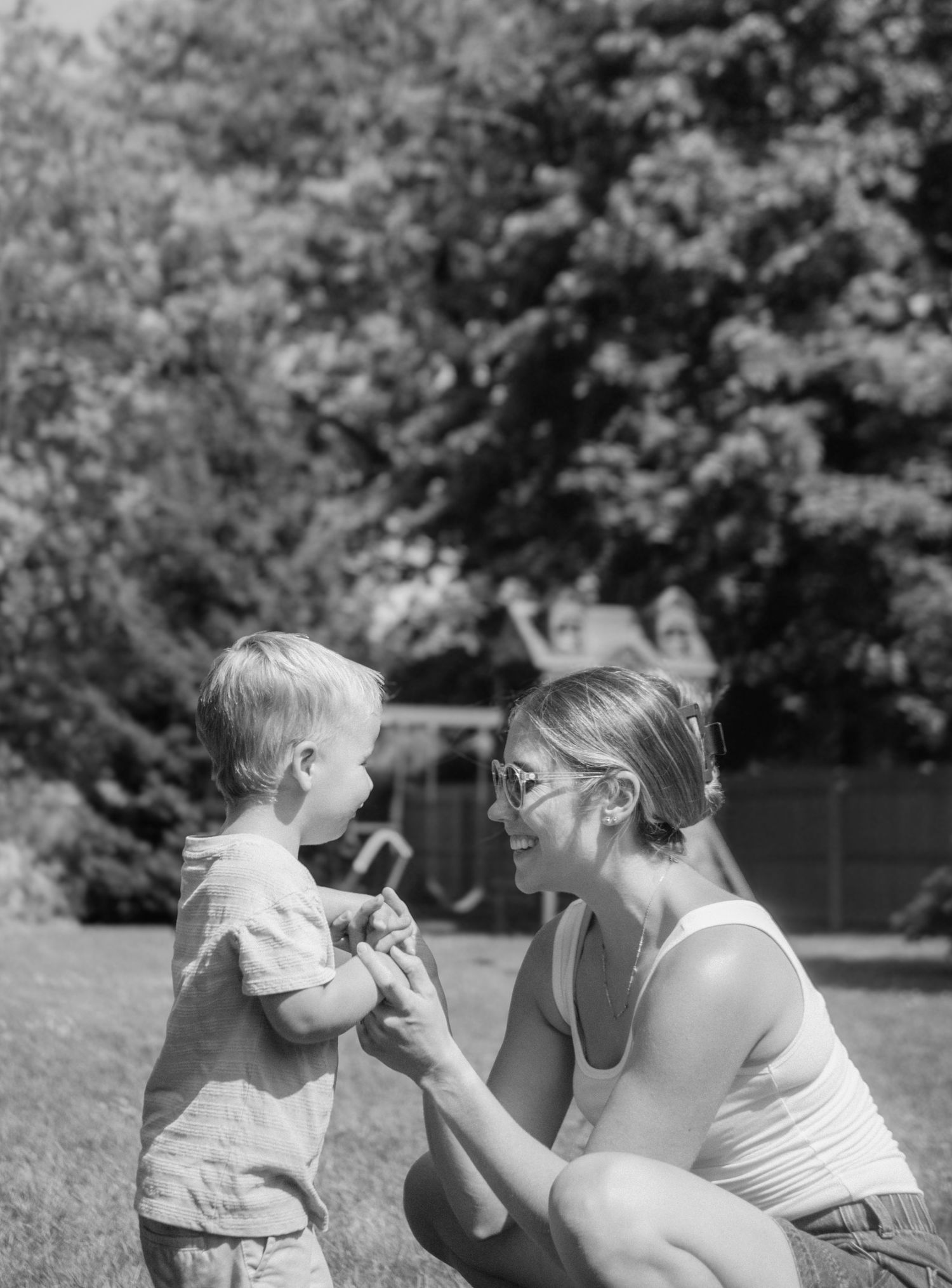 A black and white photo of a mother crouching to interact with her toddler outdoors.