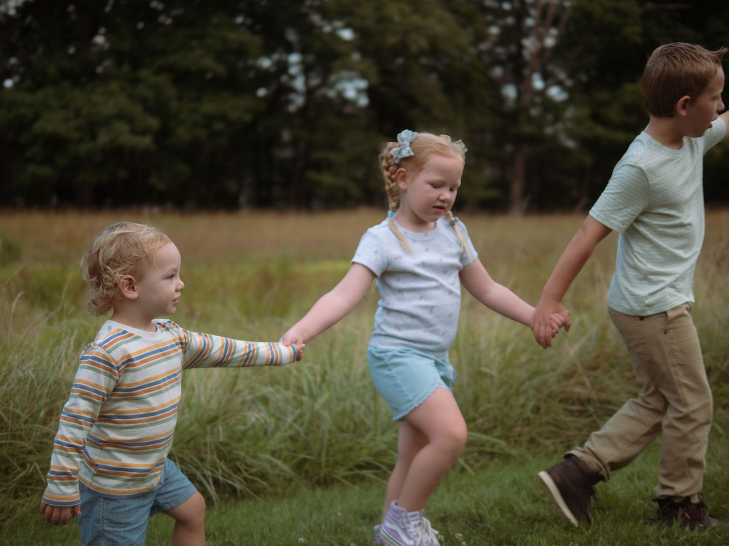Three young children holding hands and walking together in a grassy field.