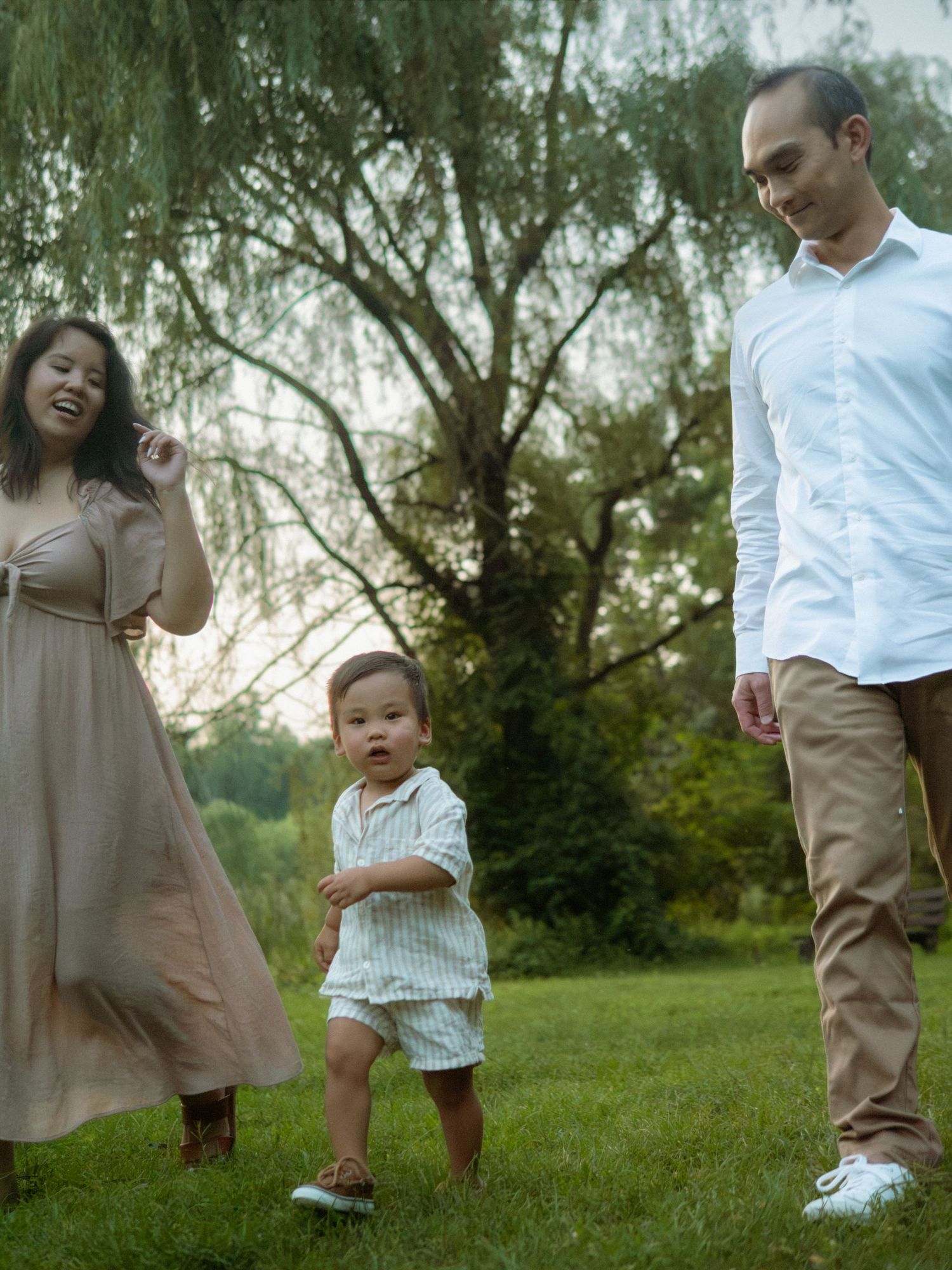 Happy family of three walking together on a grassy park lawn at dusk.