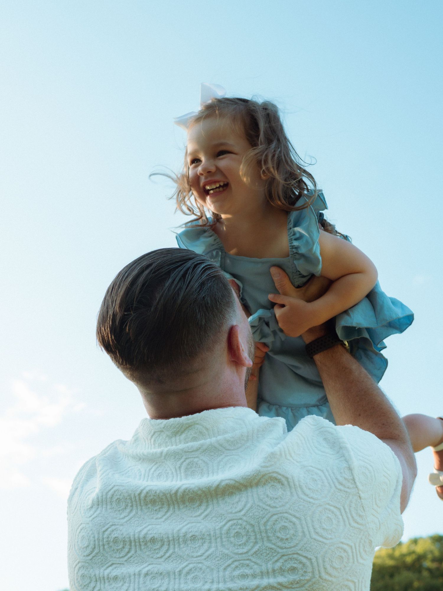 Father lifting laughing little girl in blue dress against a bright sky.