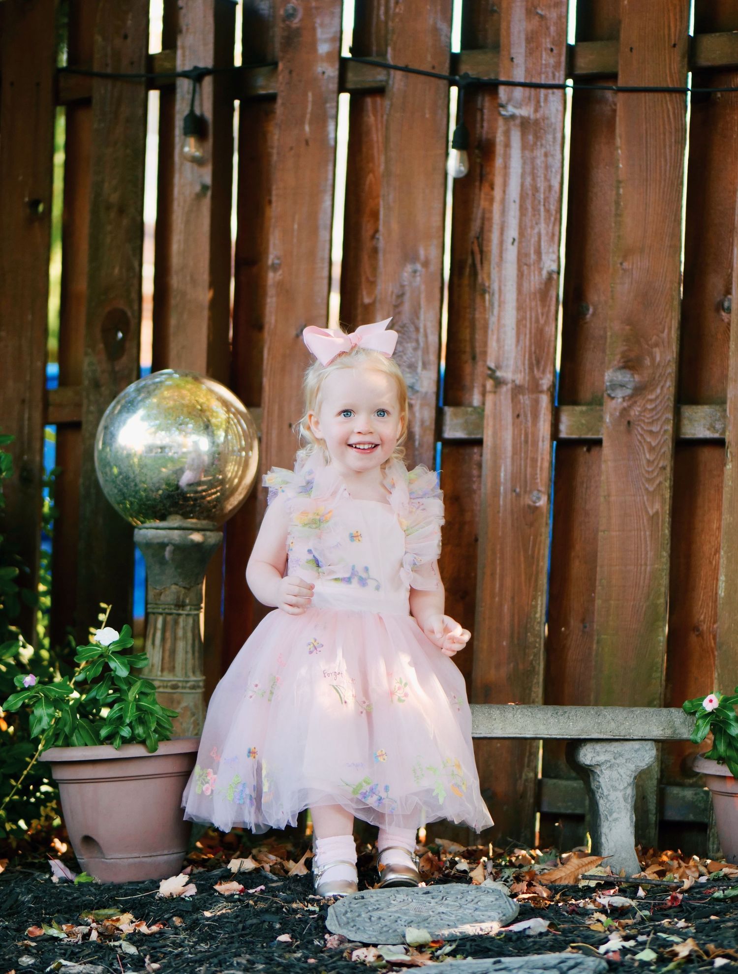 Smiling toddler girl in pink tutu dress and bow poses in garden by wooden fence.