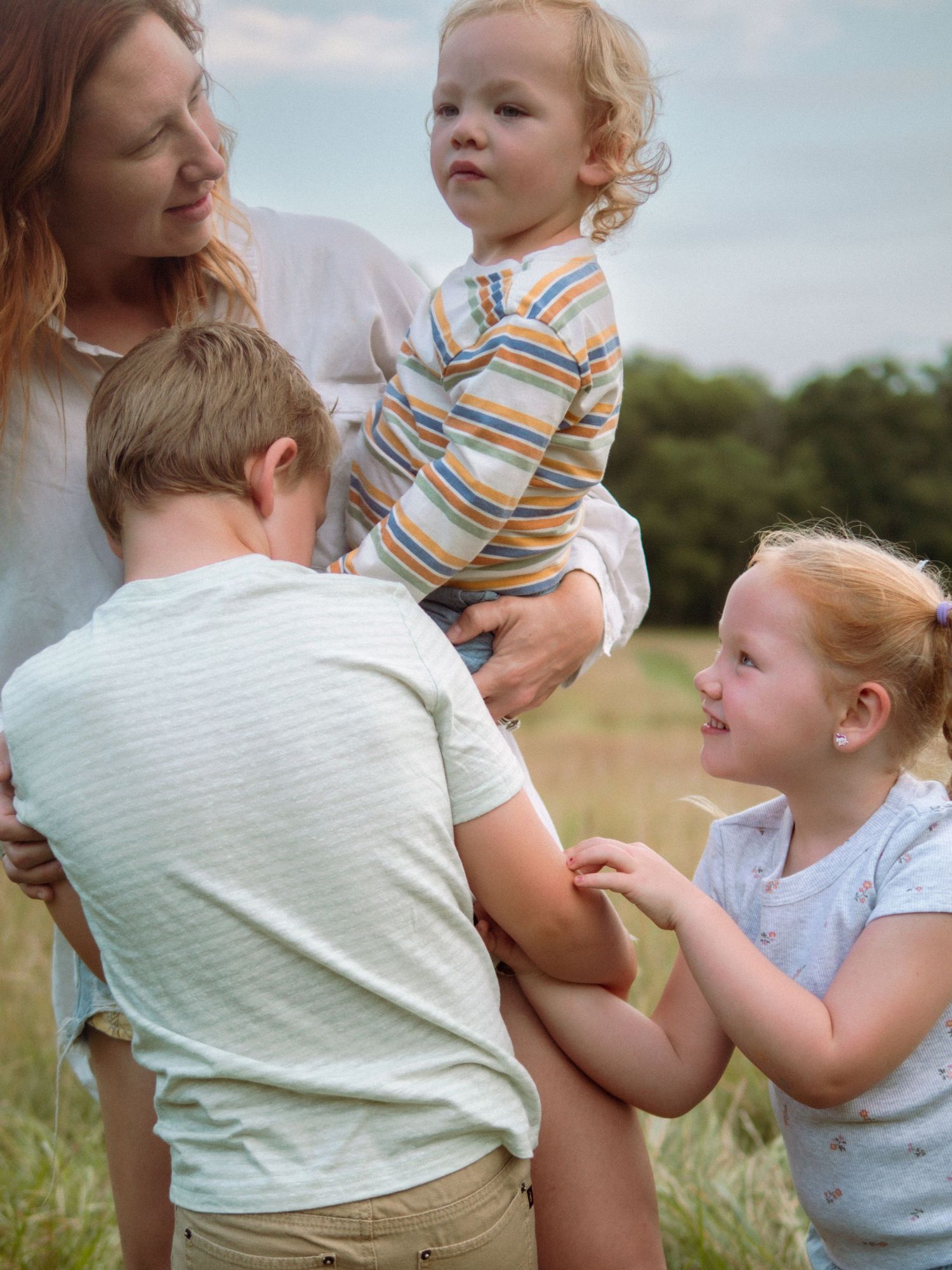 Mother in white shirt holds toddler boy while two children interact in a sunny field.