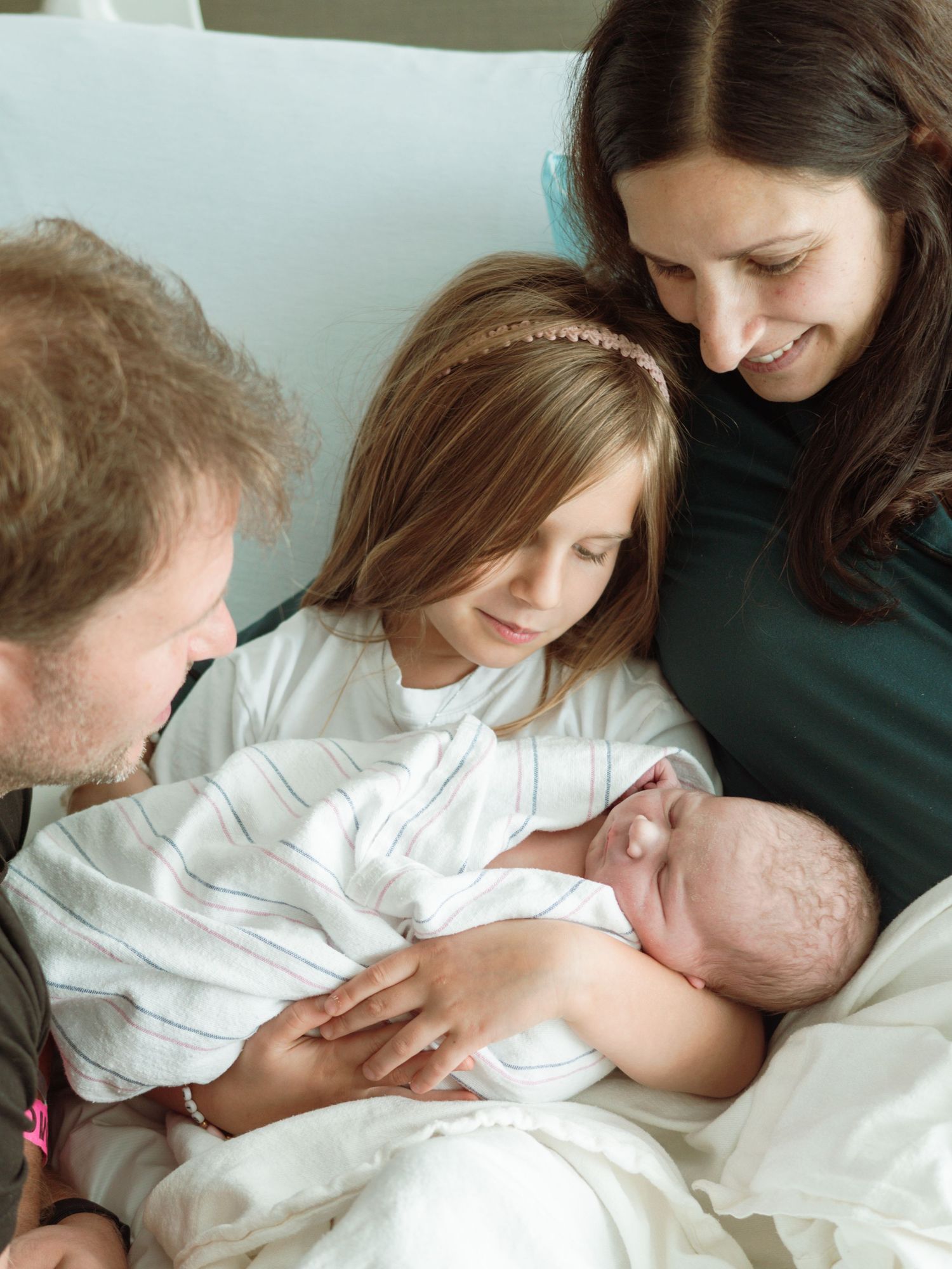 New family of four admires swaddled newborn baby together in hospital bed during fresh 48 session.