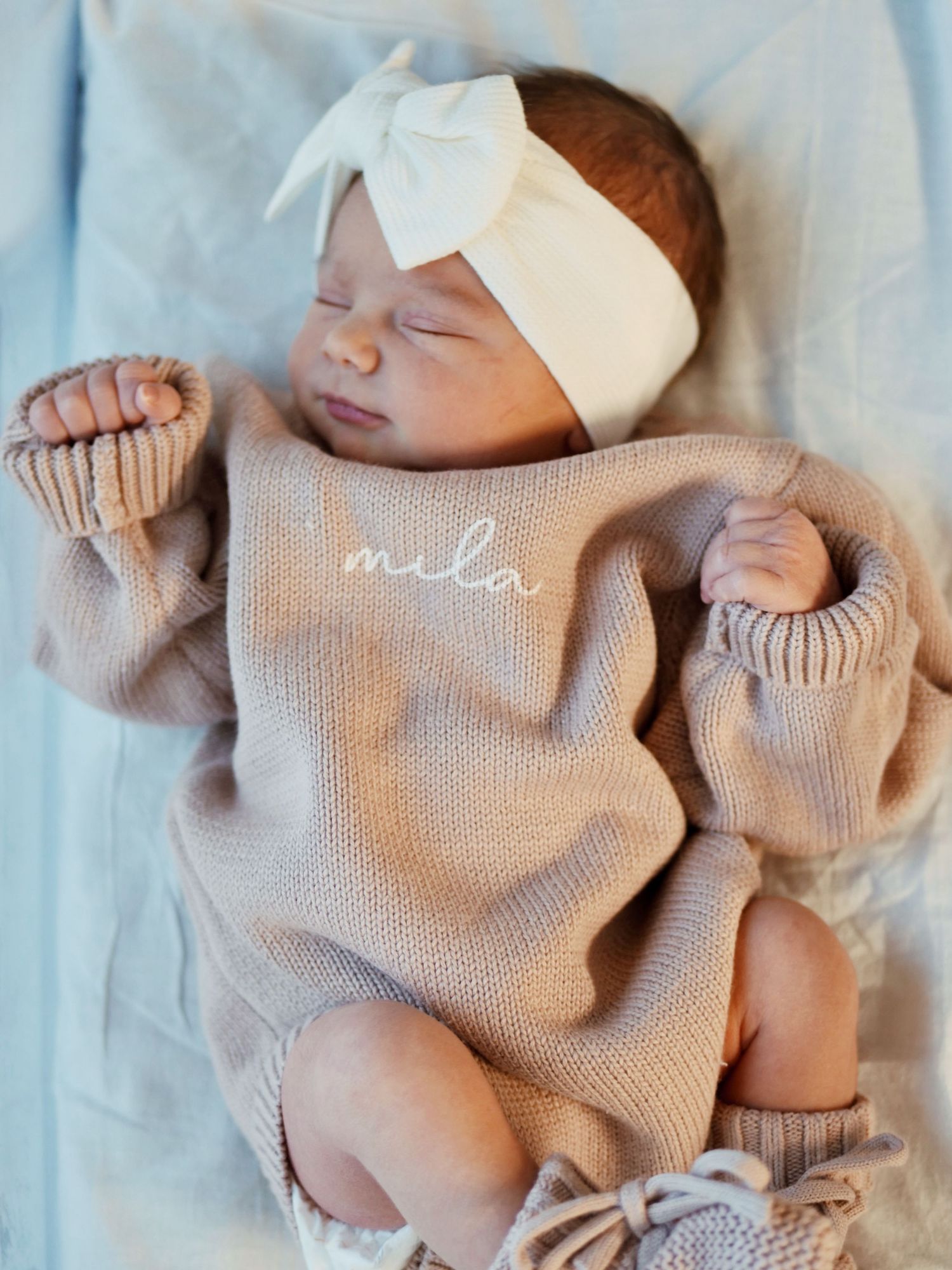 Newborn baby girl sleeping peacefully in beige knit romper with name Mila embroidered, wearing white bow headband.