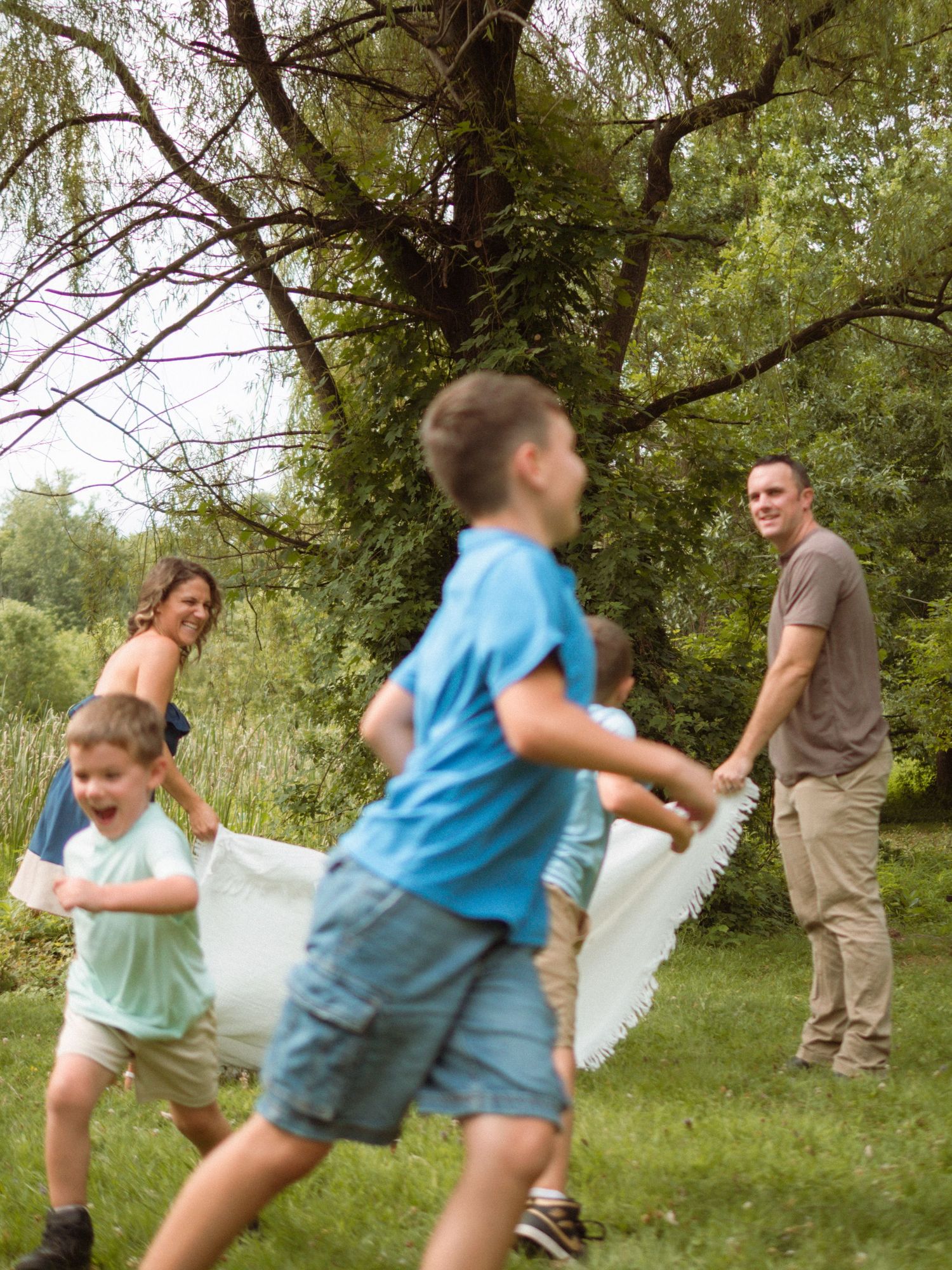 Family playing outdoors with a blanket near trees on a green lawn.
