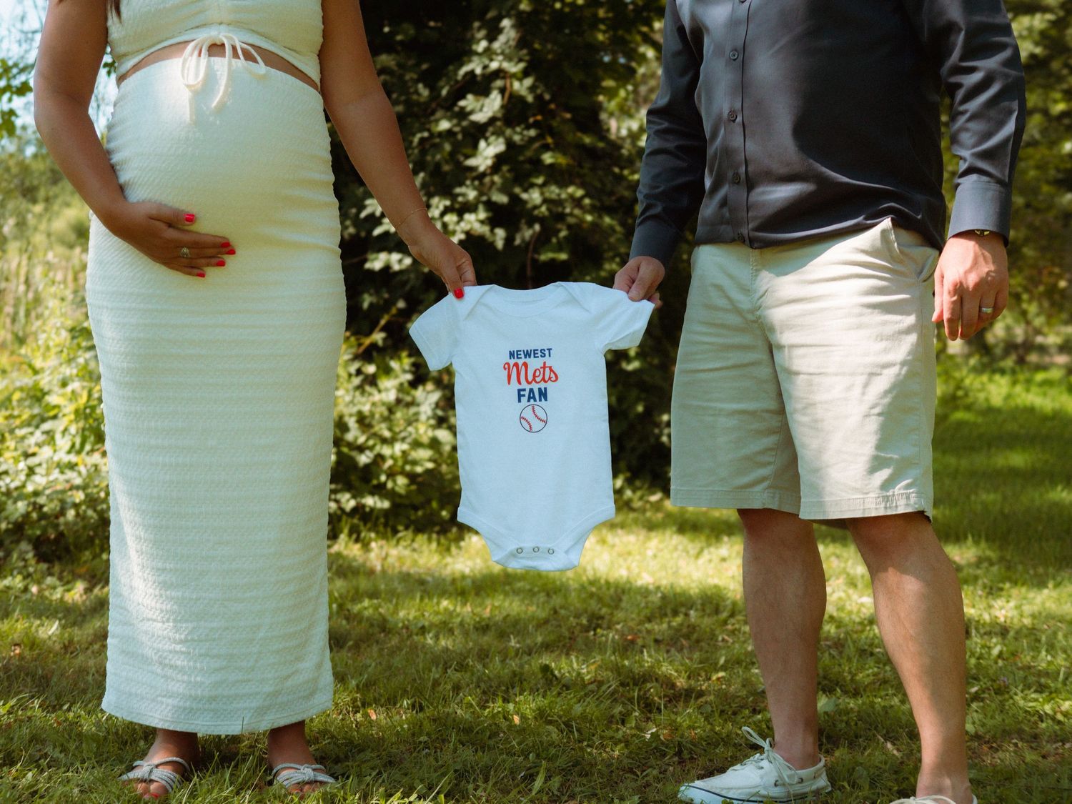 Expecting couple holding tiny baby onesie between them while standing on green lawn.
