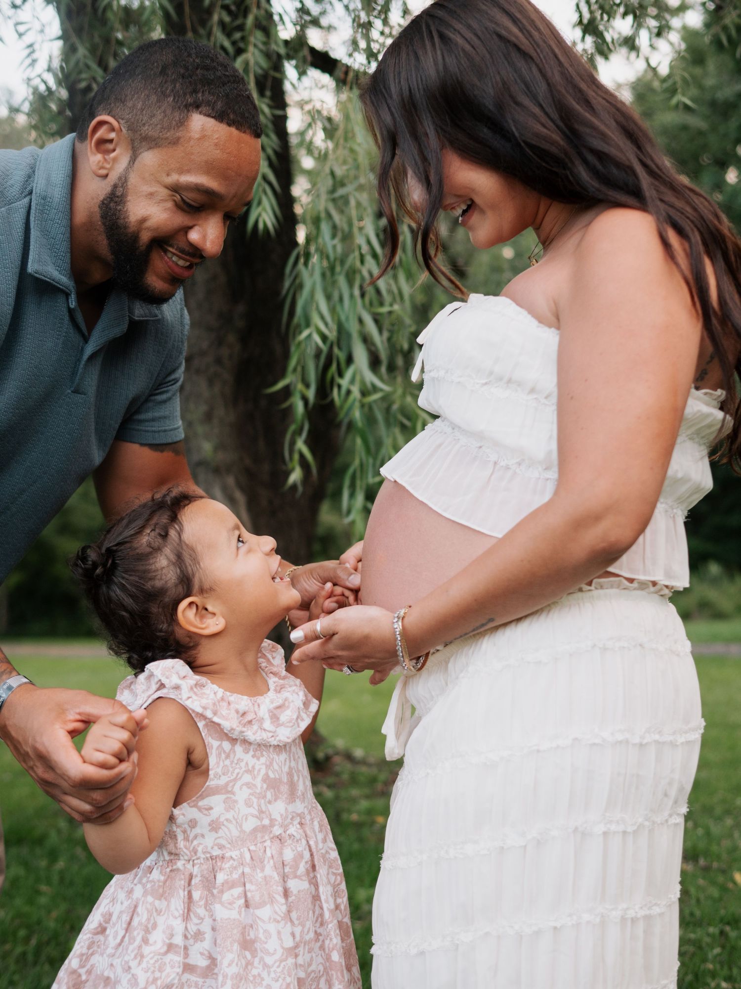 Expectant parents smiling at toddler daughter touching mothers pregnant belly outdoors near willow tree.