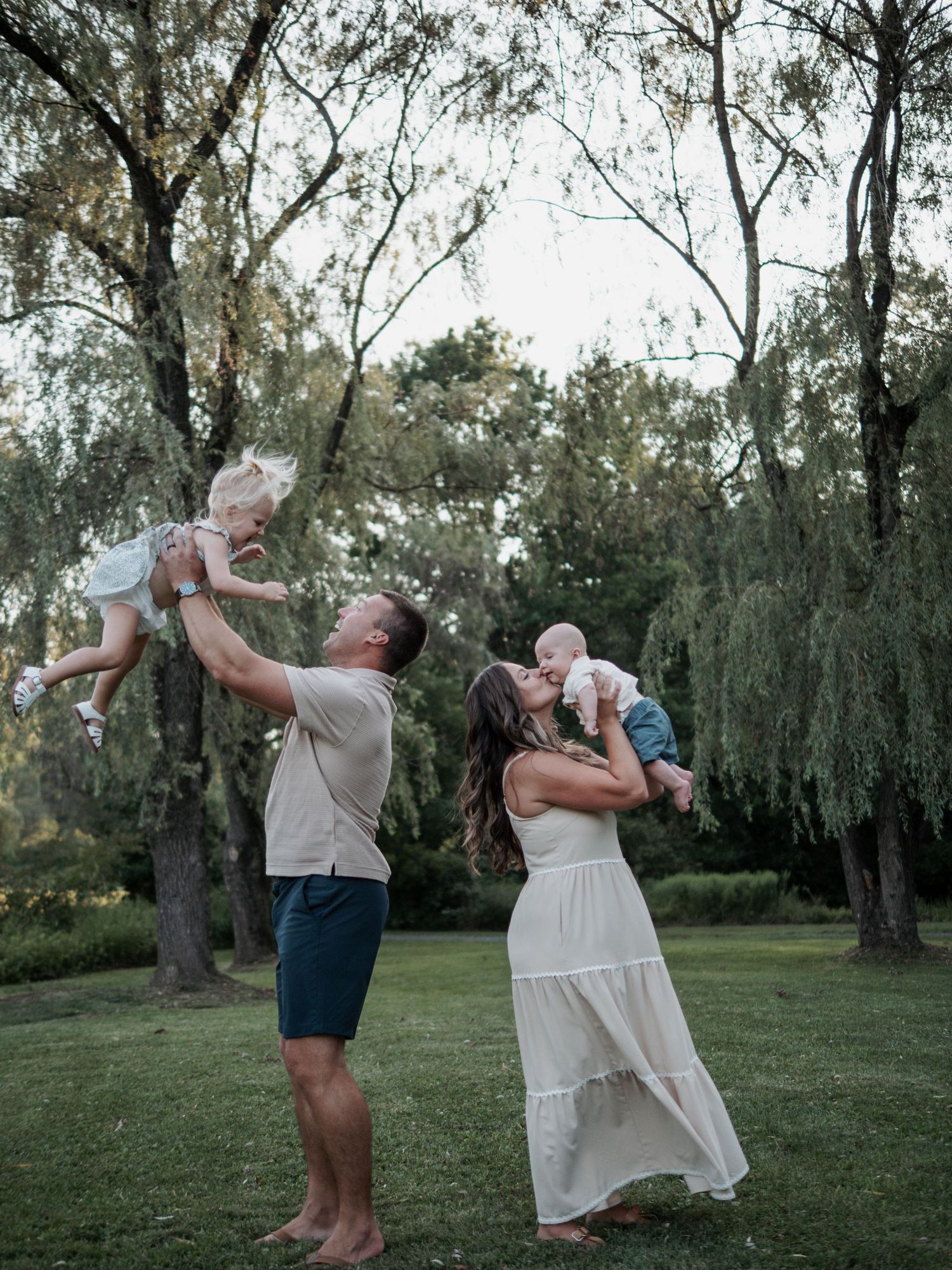 Family of four with parents lifting two young children in a lush park setting.