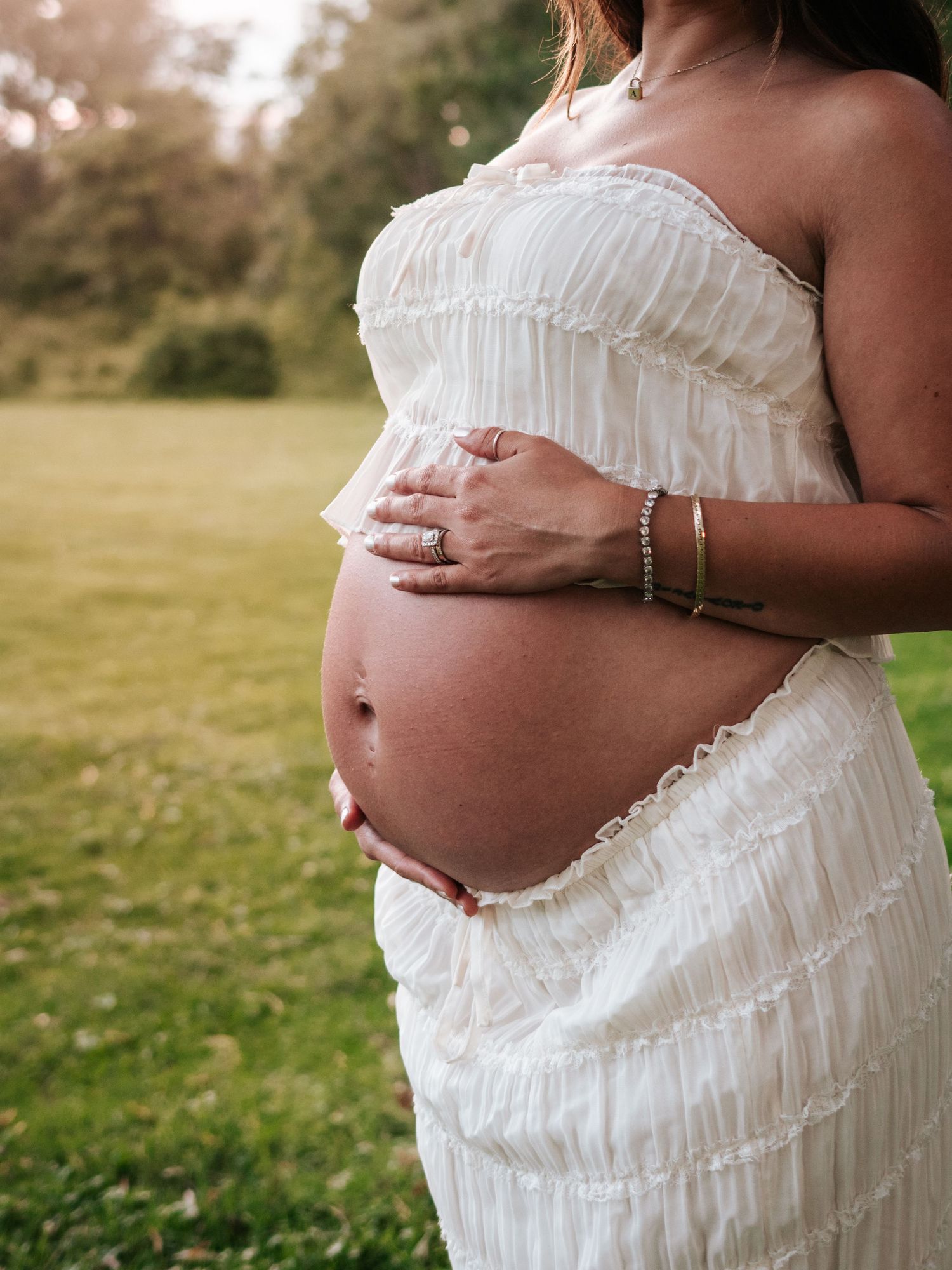 Pregnant woman in white ruffled dress cradling baby bump with jewelry-adorned hands in outdoor setting.