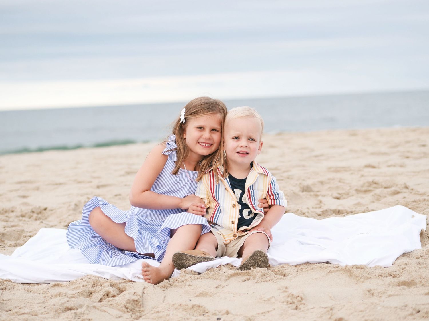 Two young siblings smiling together while sitting on a white blanket on the beach.