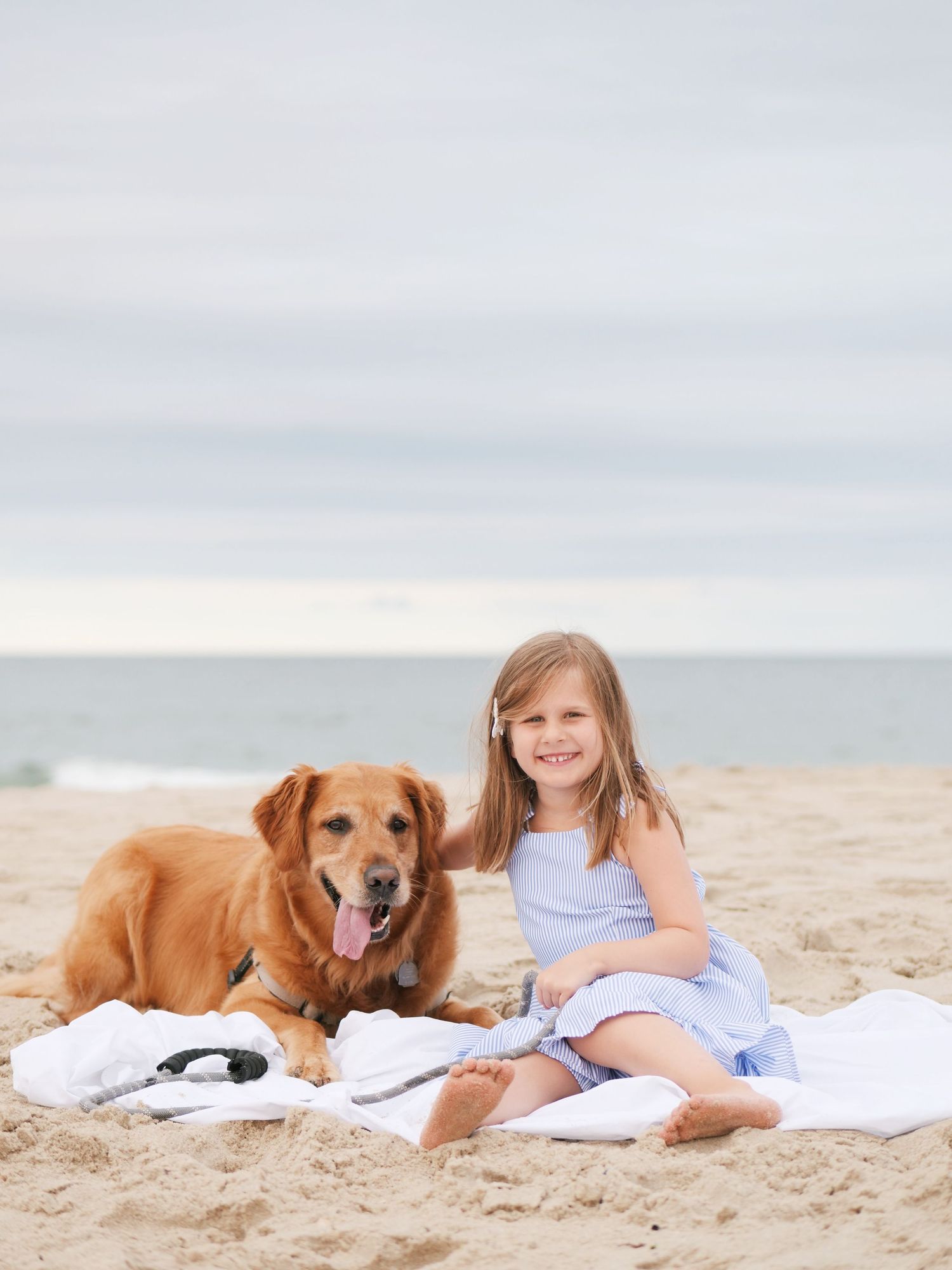 Smiling young girl in blue dress sitting on beach blanket with golden retriever dog.
