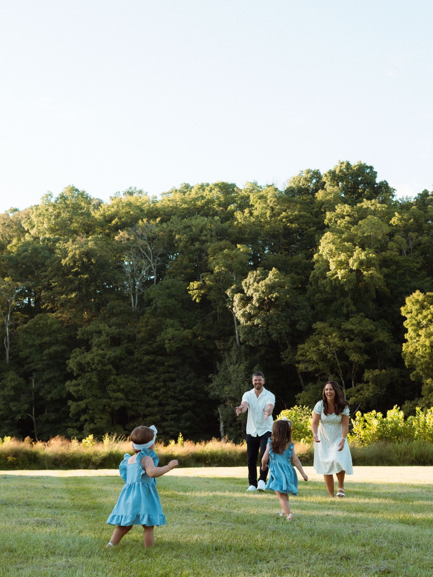 Family of four playing in a sunny green meadow surrounded by lush trees during golden hour.