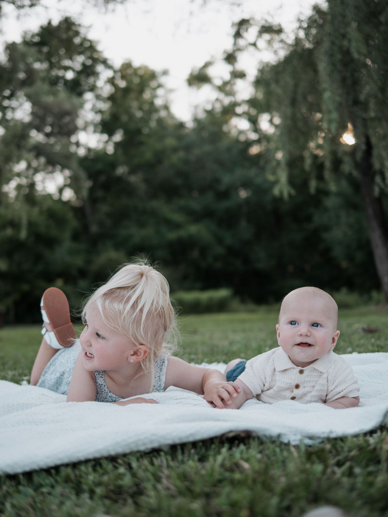 Toddler girl and baby sibling lying on white blanket on green grass outdoors.