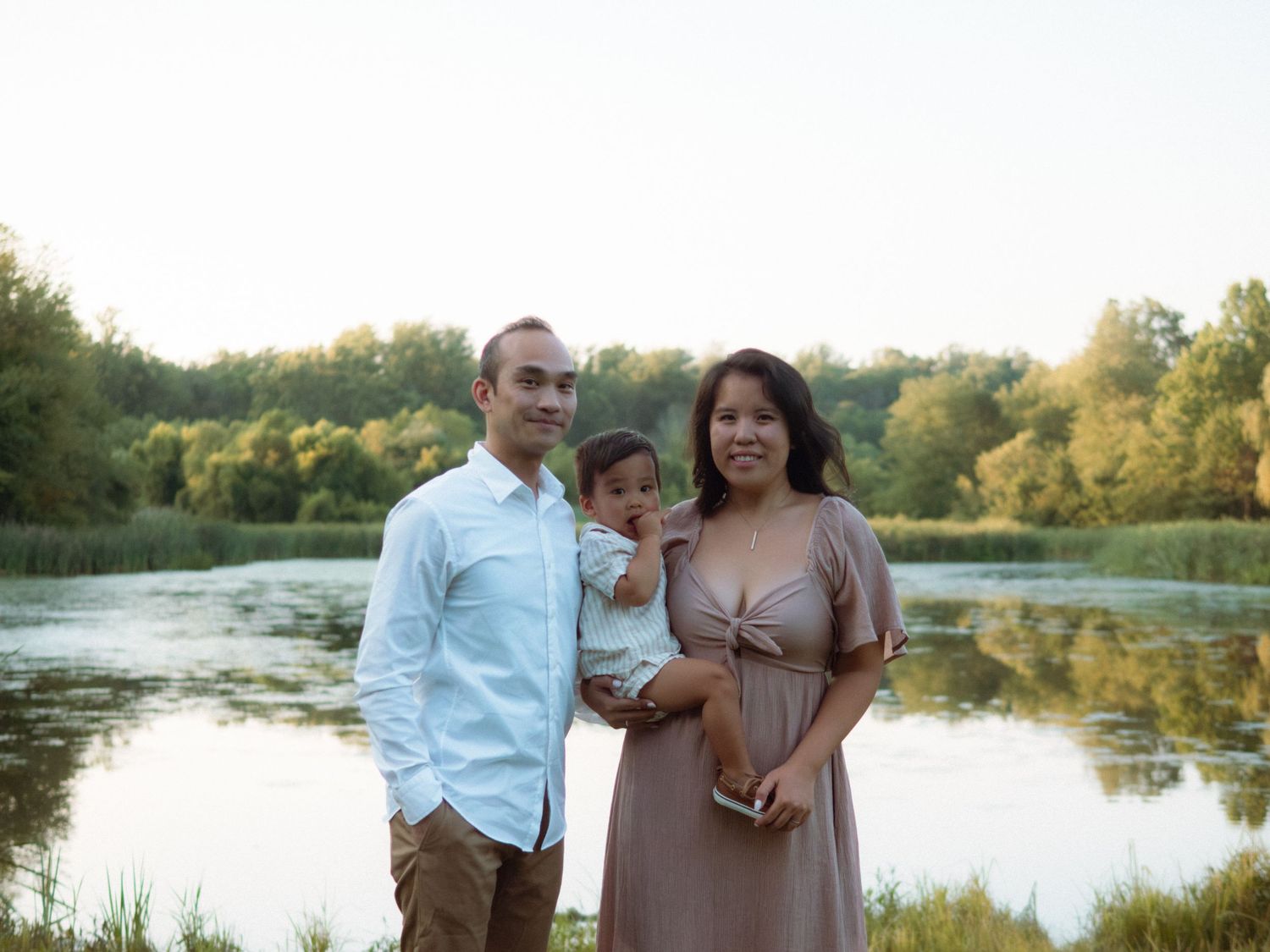 A smiling Asian family of three stands together near a serene lake at sunset.